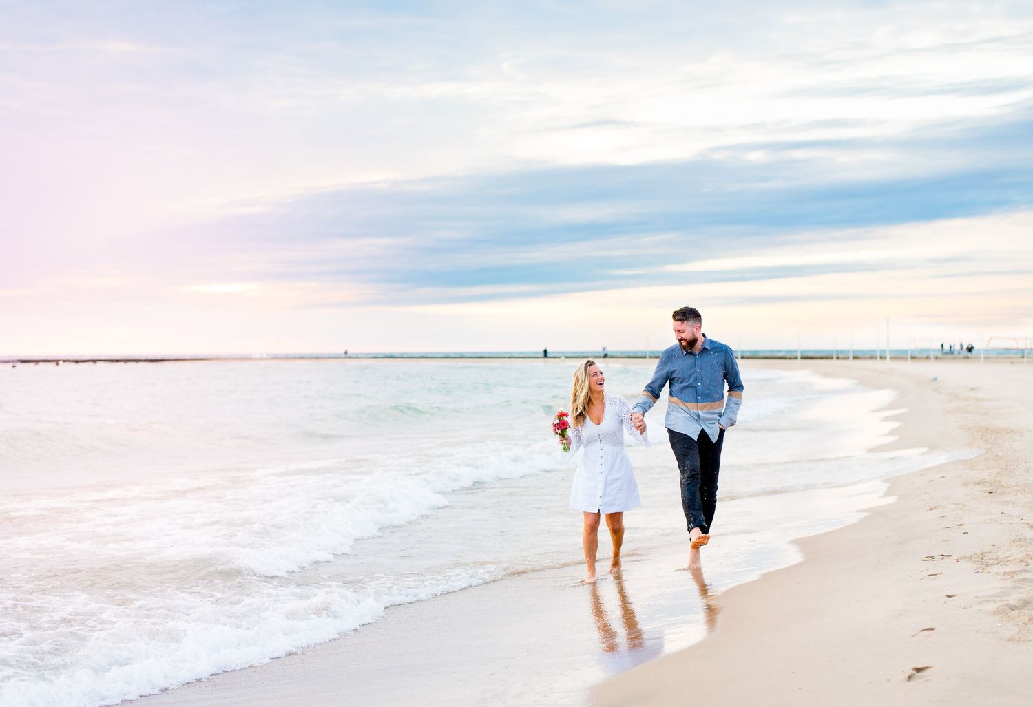 young engaged man and woman hold hands and laugh while walking along North Avenue Beach at sunrise
