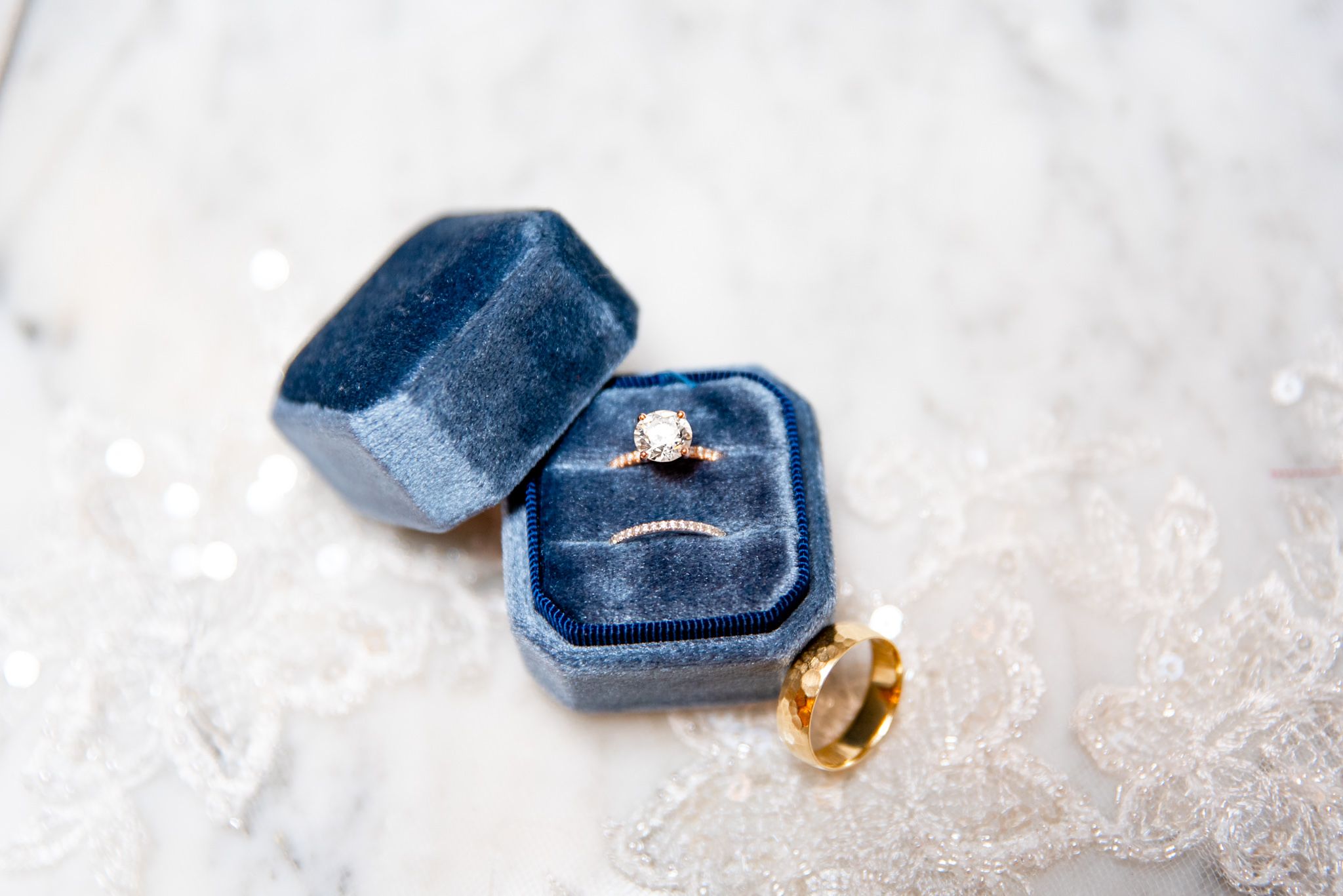 round diamond engagement ring with wedding bands in blue velvet ring box on lace wedding dress