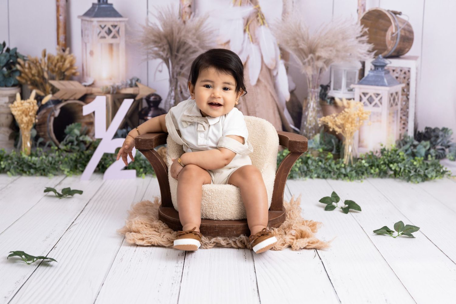 Baby sitting on cream blanket with woodland themed backdrop and decorative foliage on white wooden floor.
