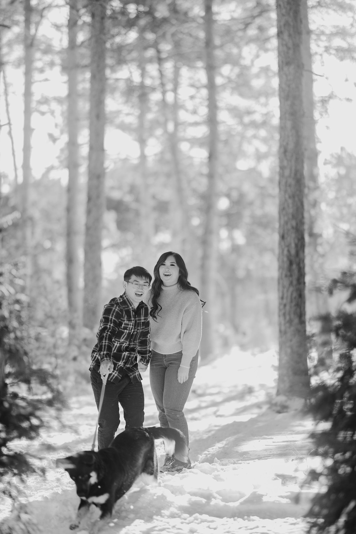Black and white photo of couple walking with dog through snowy forest trail.