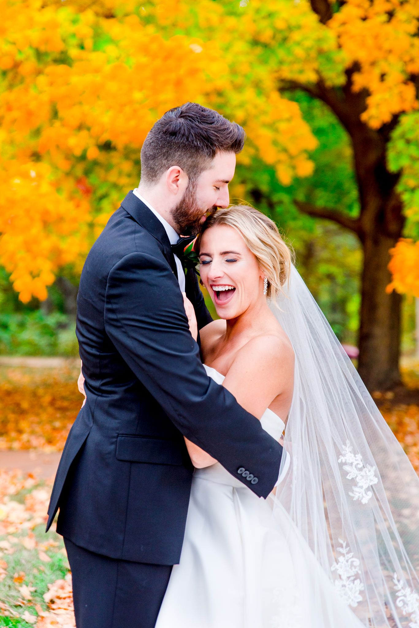 bride with lace veil leaning on grooms chest, who is hugging her in front of orange tree for STL wedding pictures