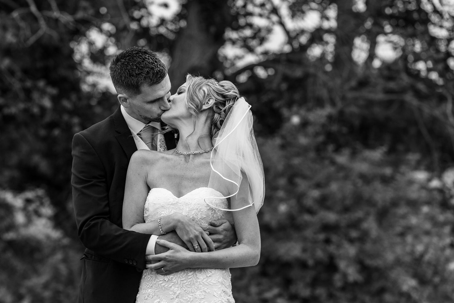 Groom stands behind the bride as she turns her head to kissing his arms around her waist