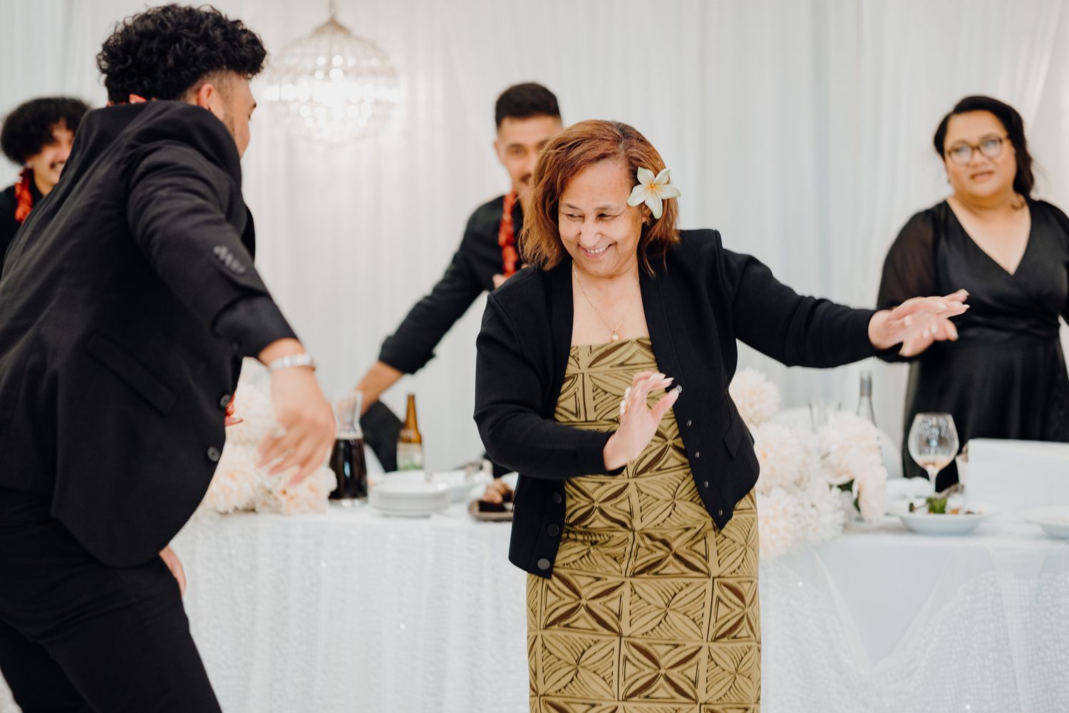 Guests dancing energetically at a wedding reception with white table settings and chandeliers in the background.