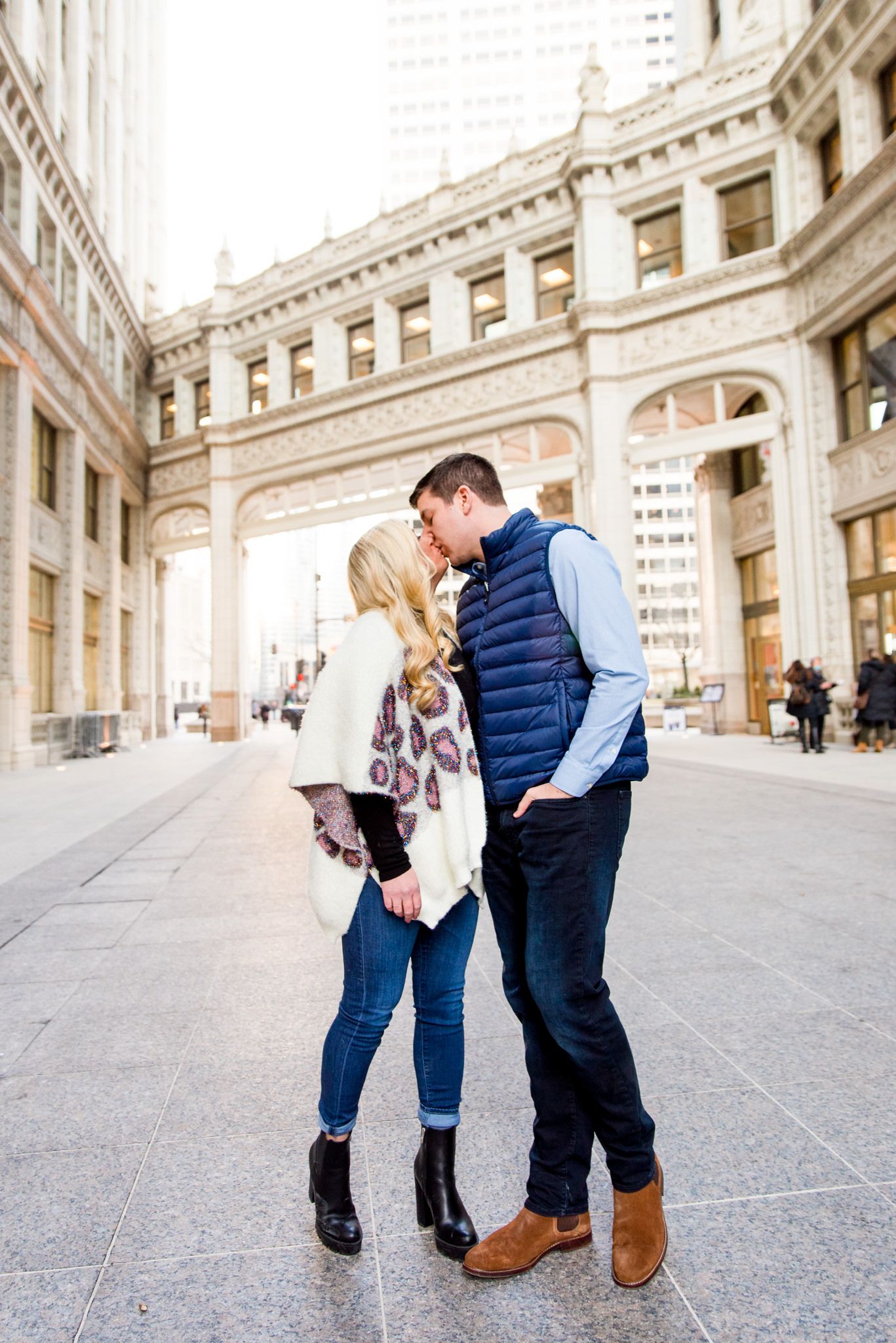 blonde woman in leopard cardigan kissing fiance in blue vest in front for Wrigley Building engagement pictures