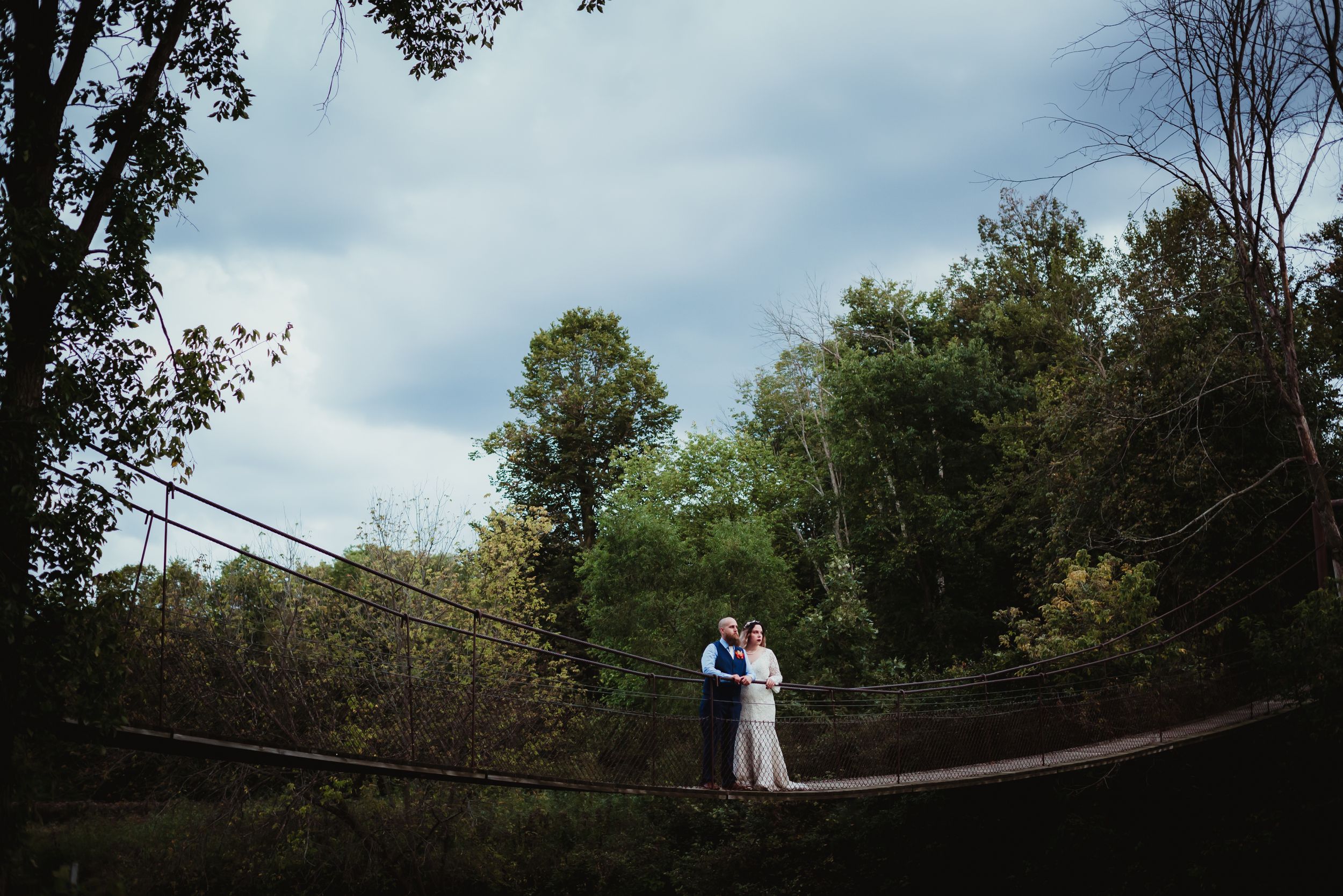 Man and woman in wedding attire standing in the middle of a swinging cable rope bridge.