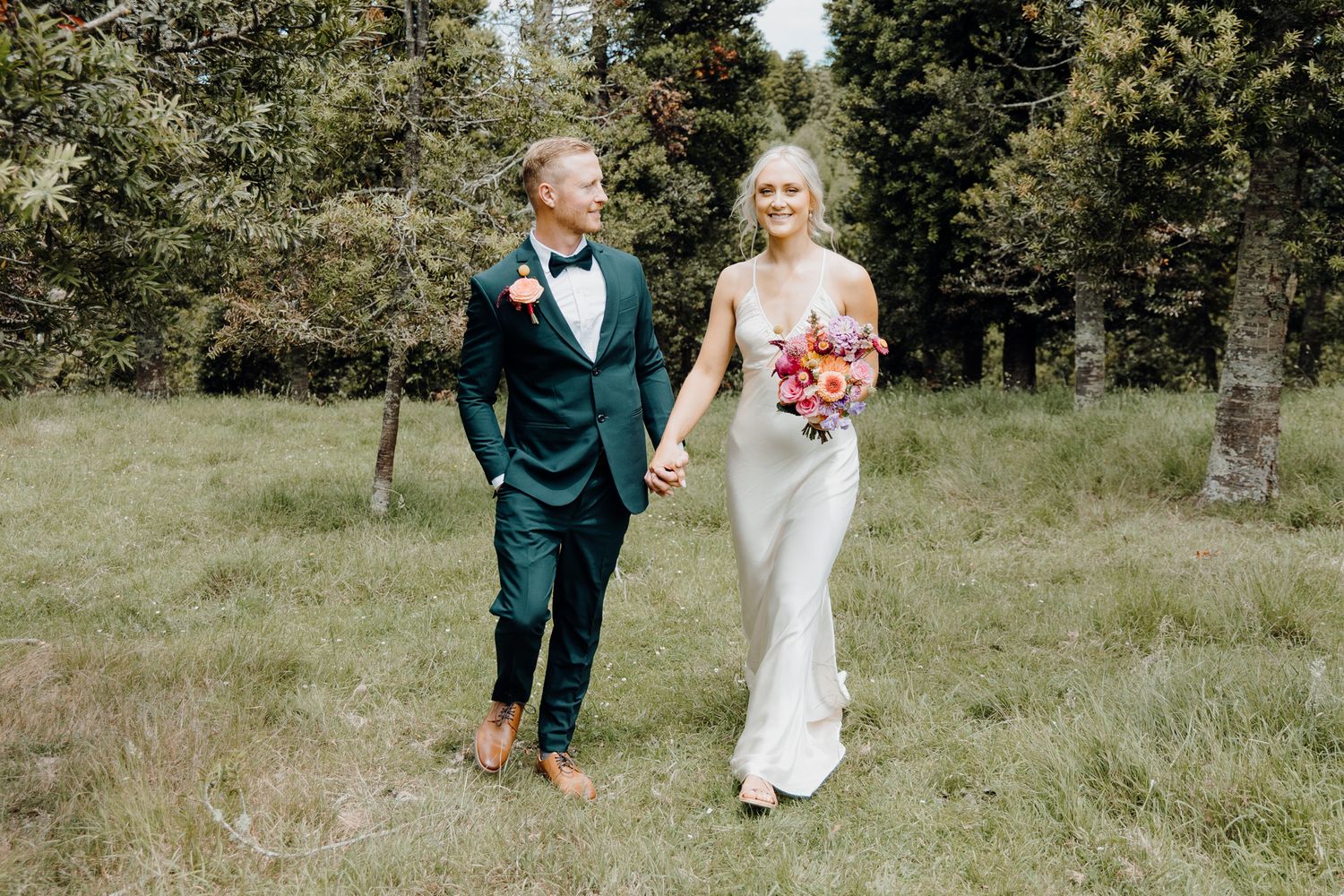 Wedding couple walking through a green field with colorful bouquet.