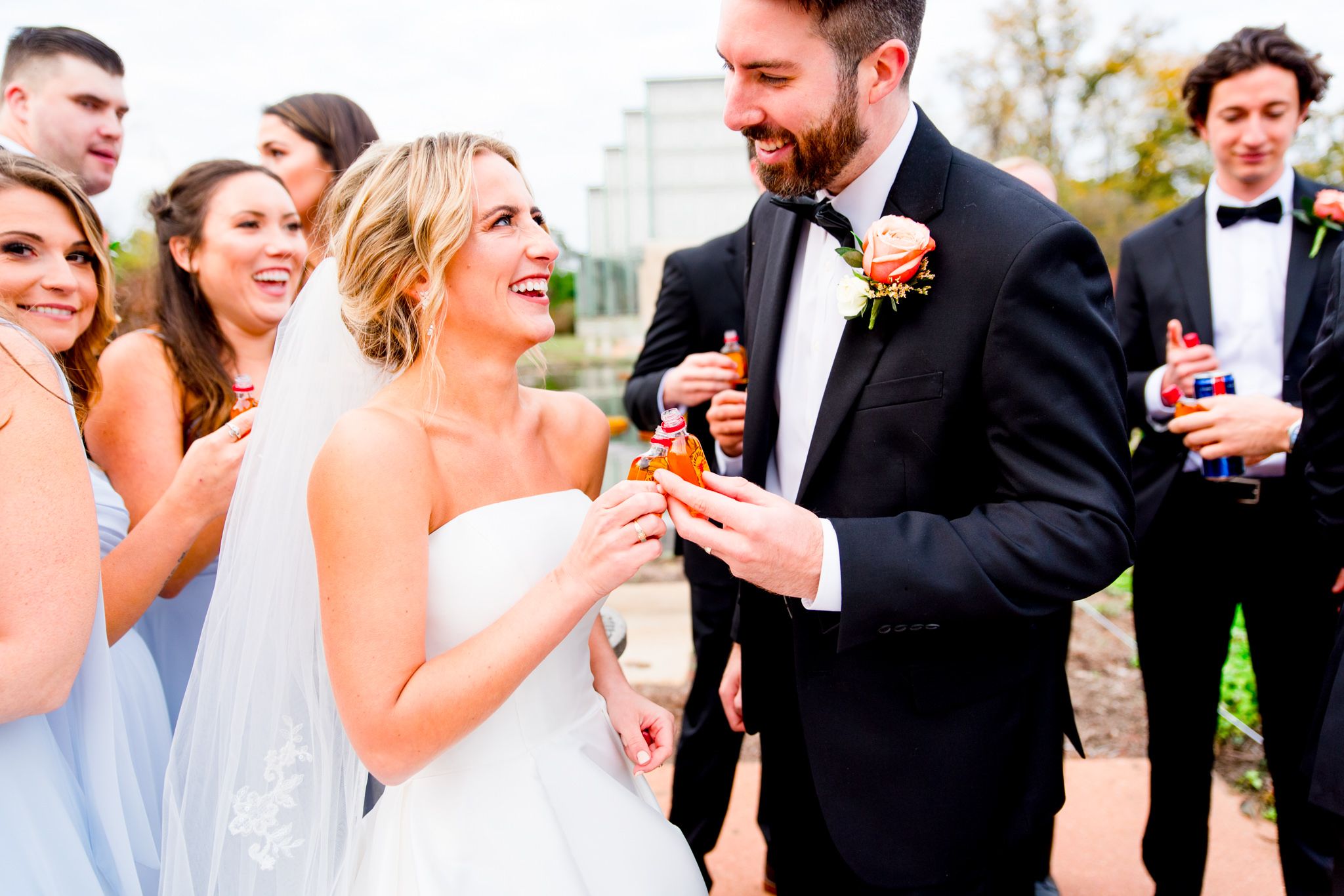 bride and groom smiling at each other, clinking mini fireball bottles together on wedding day