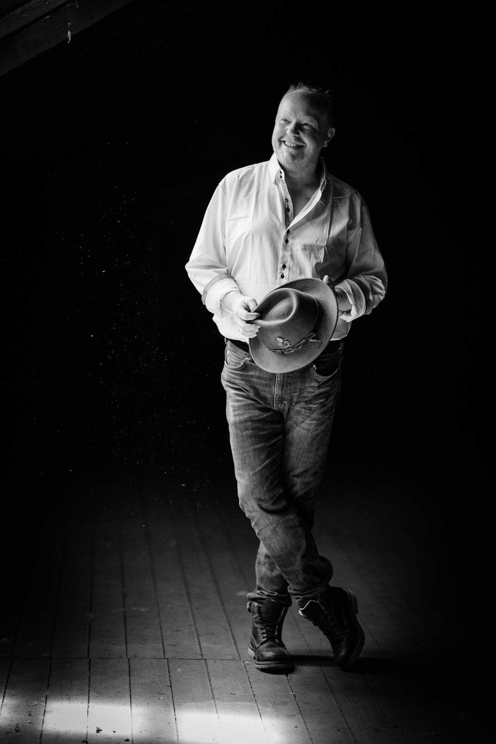 Black and white portrait of someone in a casual white shirt and jeans holding a cowboy hat while standing on wooden floor.