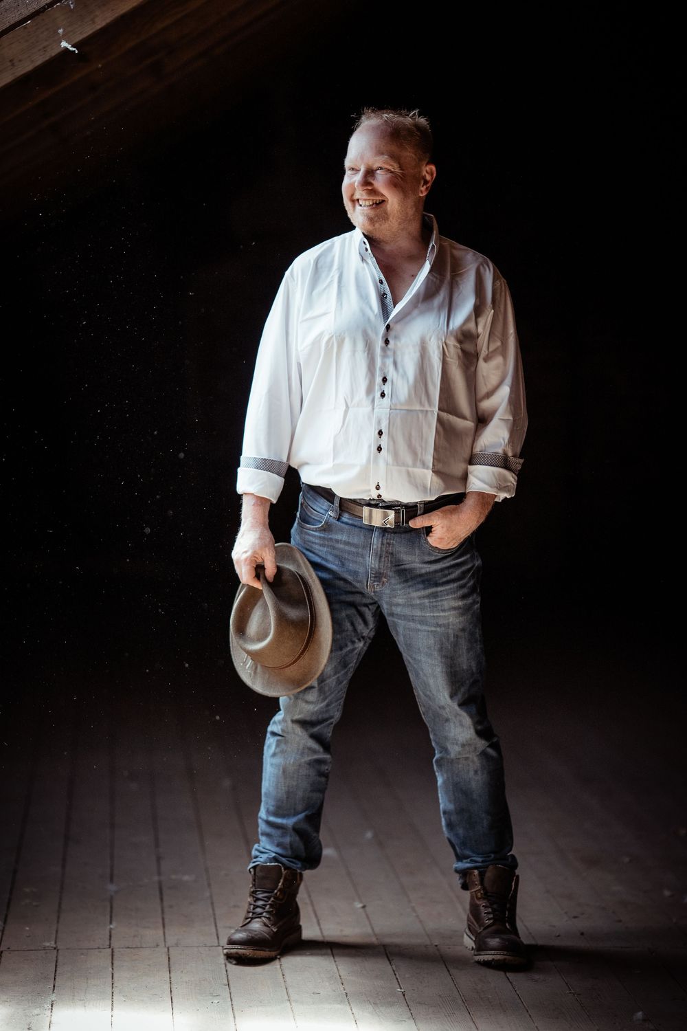 A figure in a white shirt and blue jeans stands dramatically against a dark background while holding a hat.