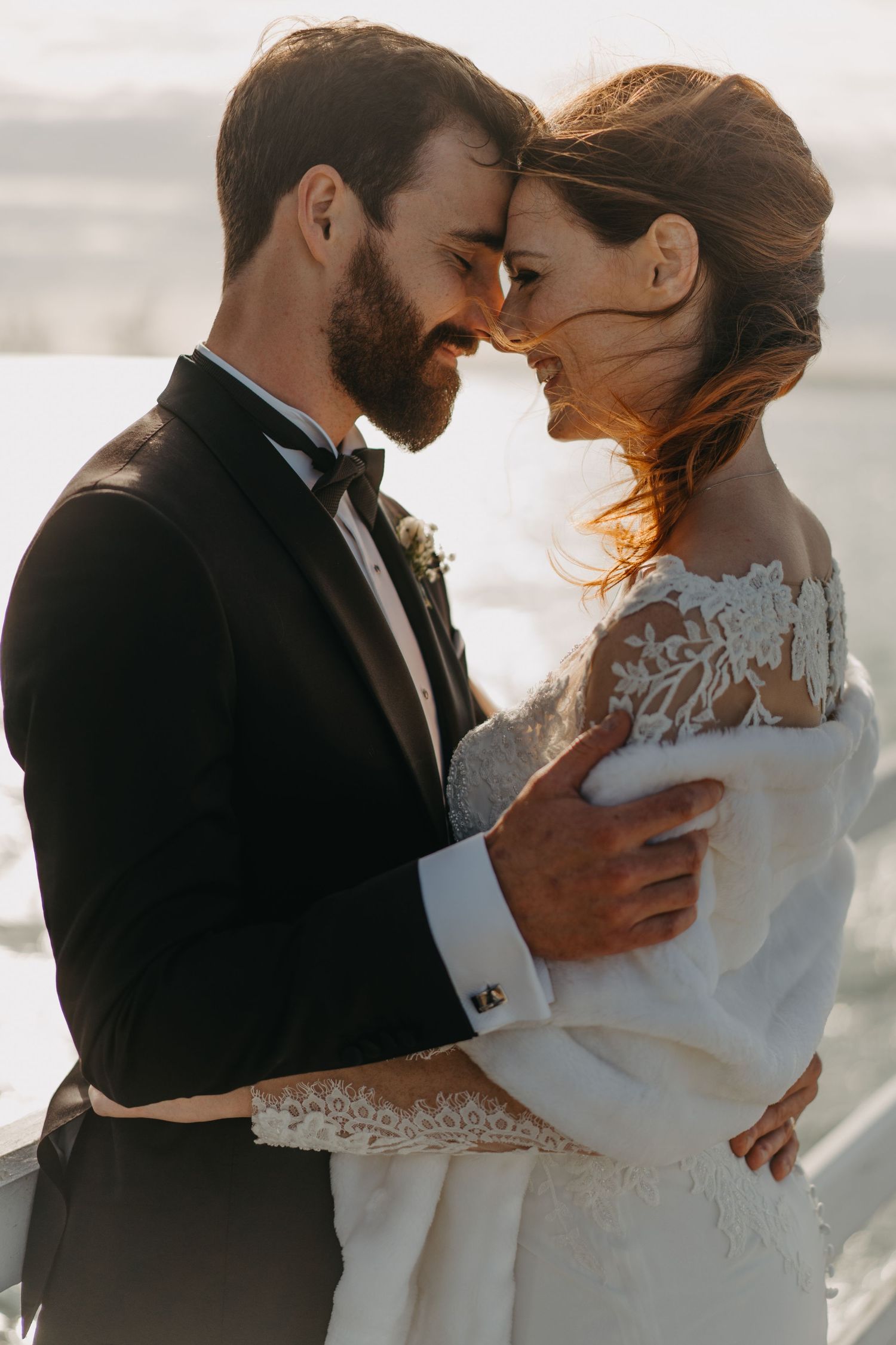A tender moment between newlyweds in formal attire against a sunlit coastal backdrop.