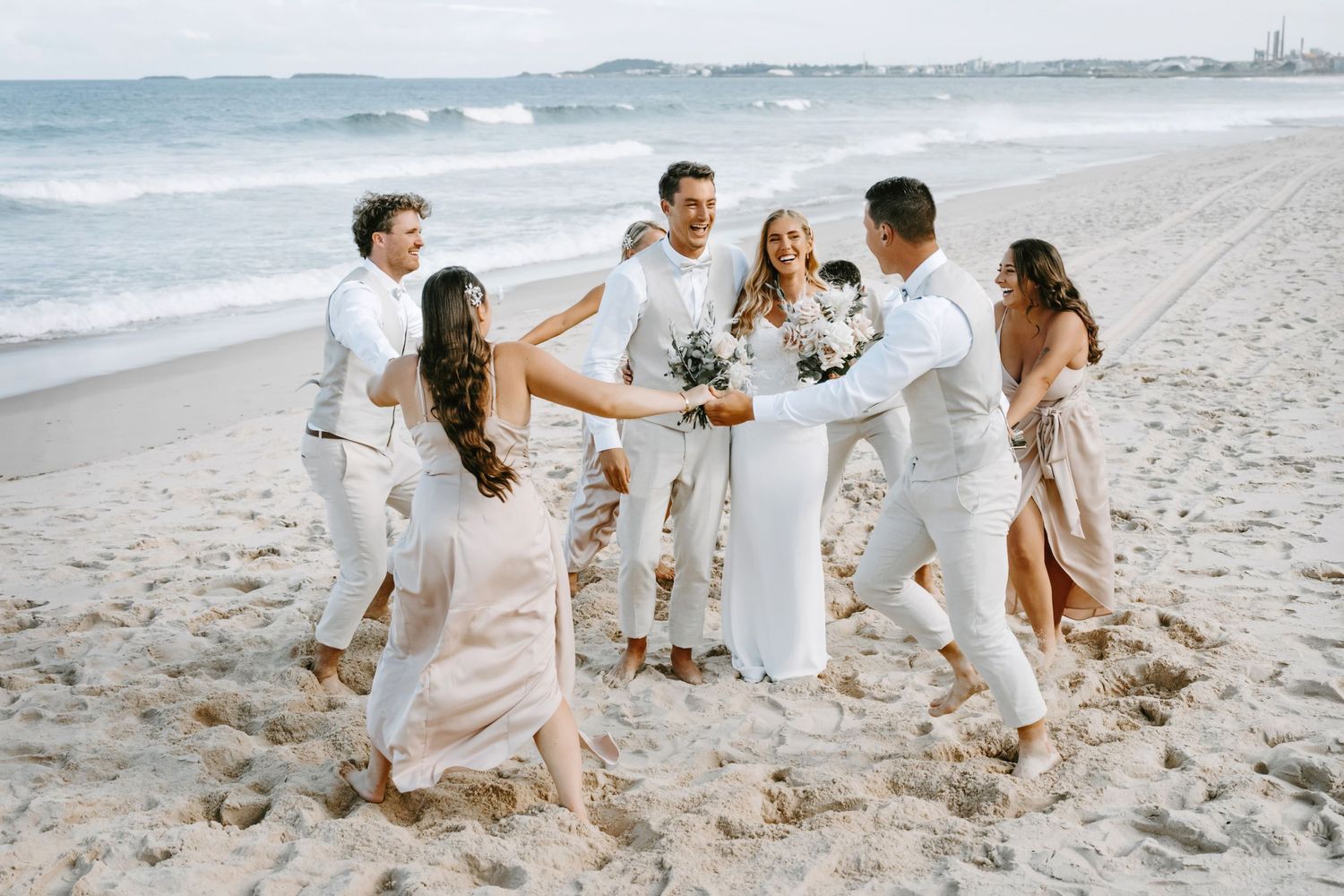 A wedding party celebrates on a beach, dressed in white and beige outfits against the ocean backdrop.