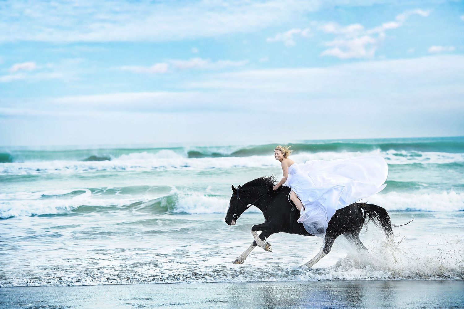 A dramatic wedding photo of someone riding a black horse through ocean waves in a white dress.