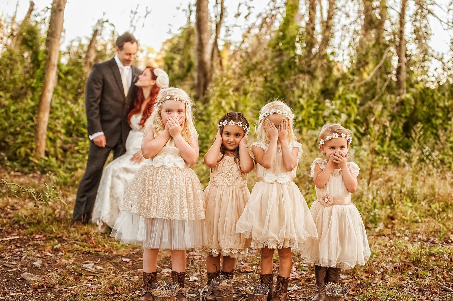 Four flower girls in lace dresses follow behind a suited figure in a wooded outdoor setting.
