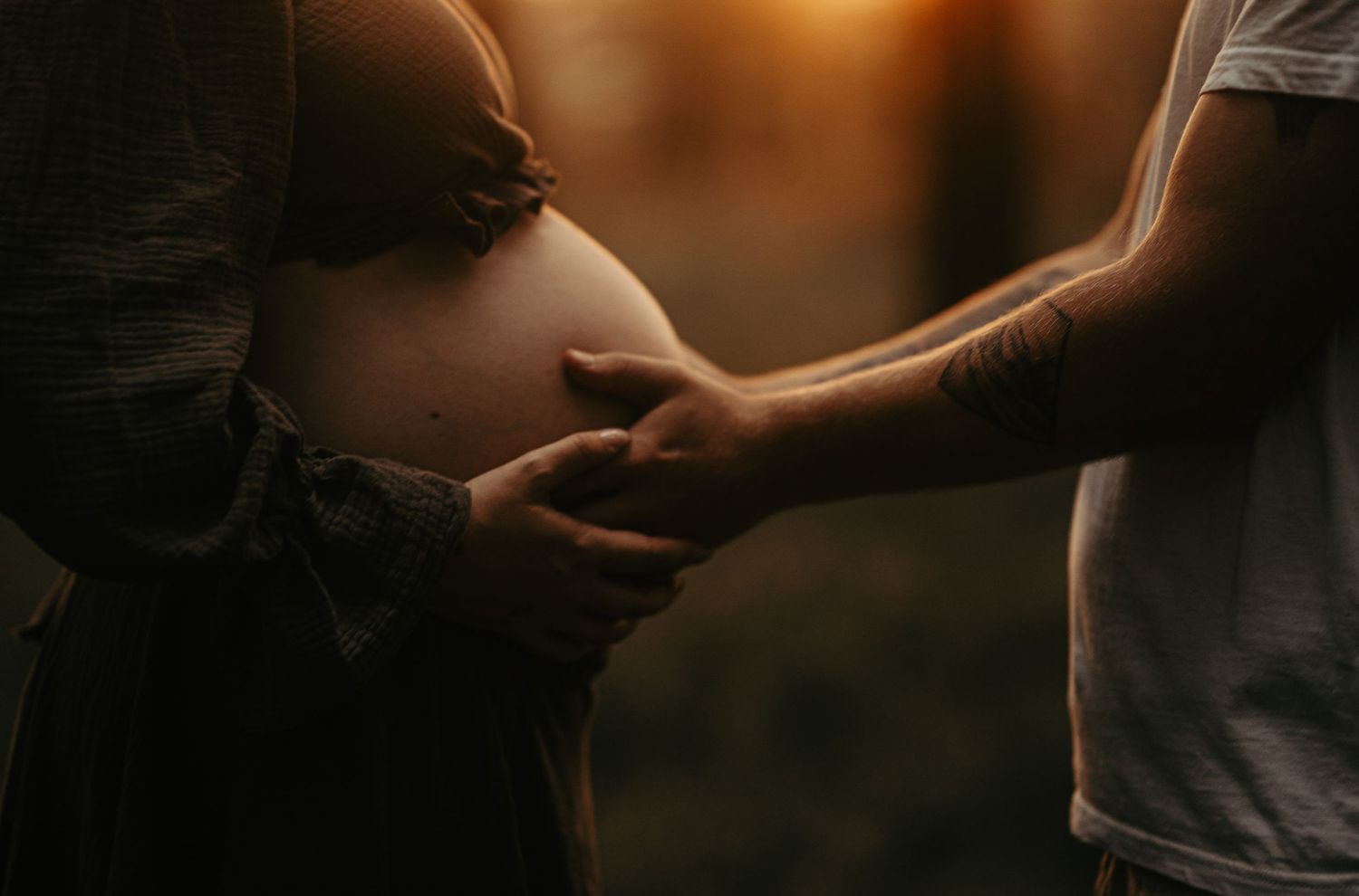Close-up silhouette of hands gently touching a pregnant belly at sunset.