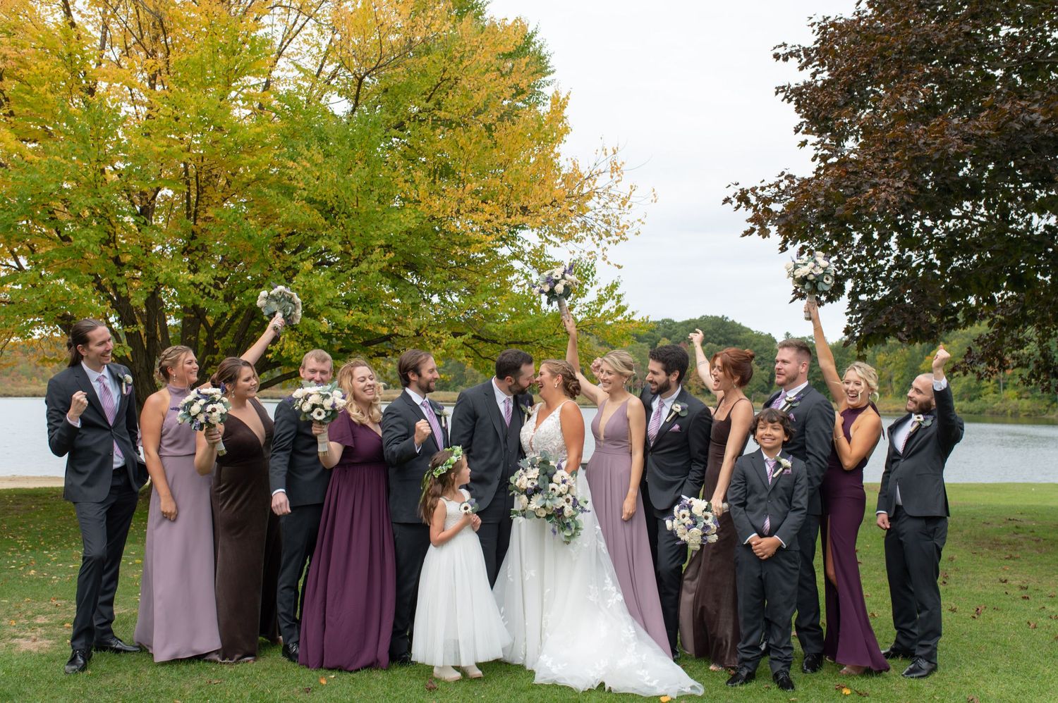 Wedding party celebrates with raised arms during group photo on lawn with autumn trees.