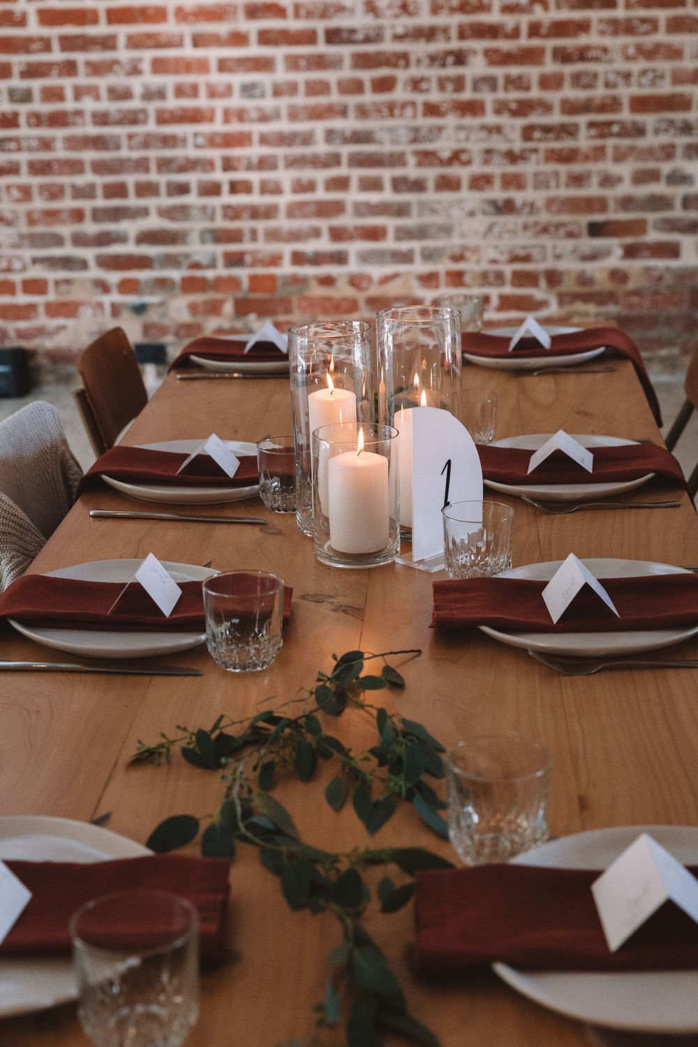 Modern wedding table setting with glass candle holders and burgundy napkins on wooden dining table.