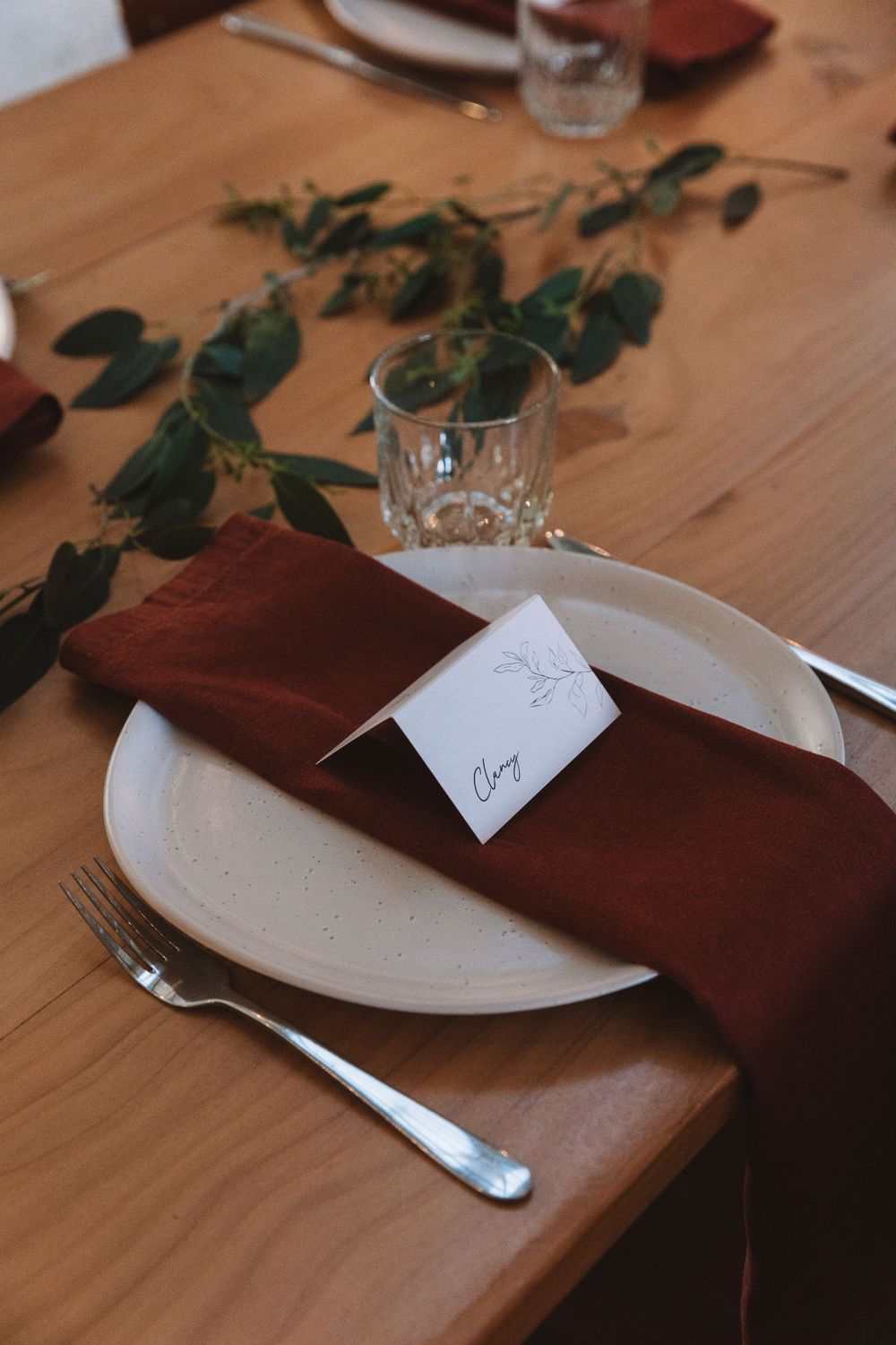 Individual place setting with burgundy napkin, white plate, and greenery accents on wooden table.