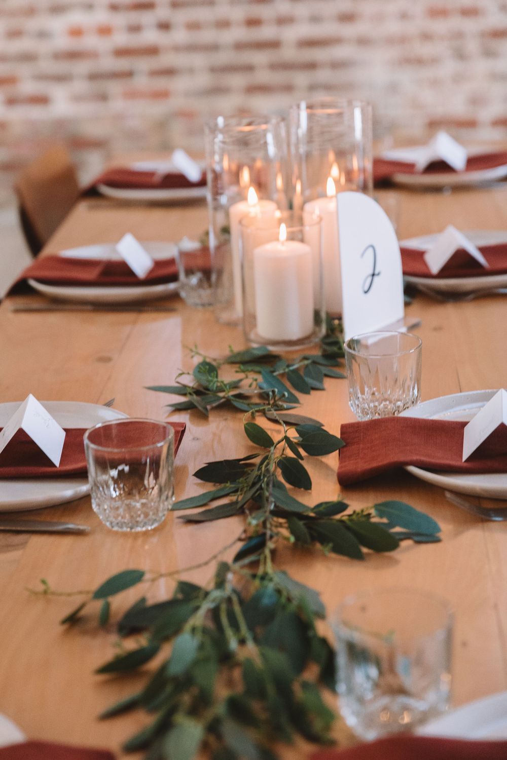 Elegant table setting with white candles, greenery, and burgundy napkins against exposed brick wall.