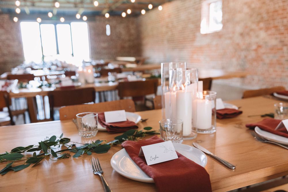 Industrial venue with wooden tables set for dining featuring white candles and burgundy accents.