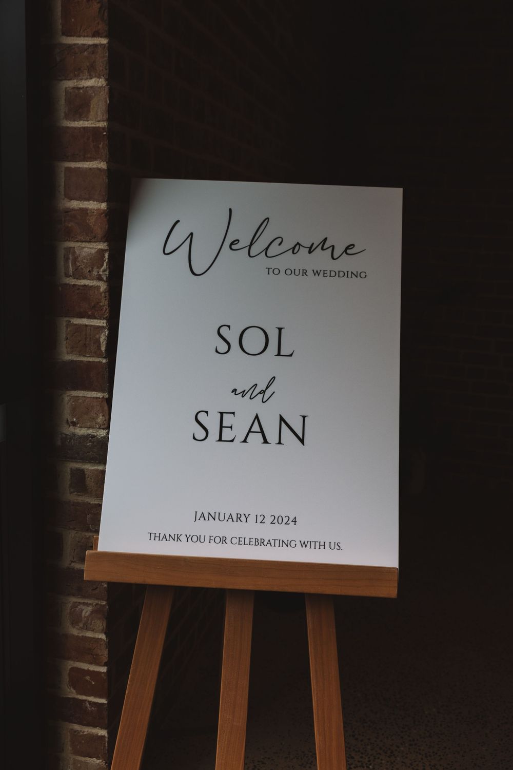 Welcome sign for Sol and Sean's wedding on January 12 2024 displayed on wooden easel against brick wall.