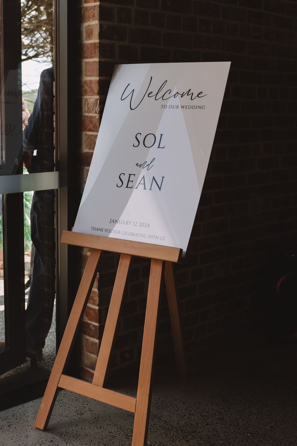 Wedding welcome sign on wooden easel against dark brick exterior wall.