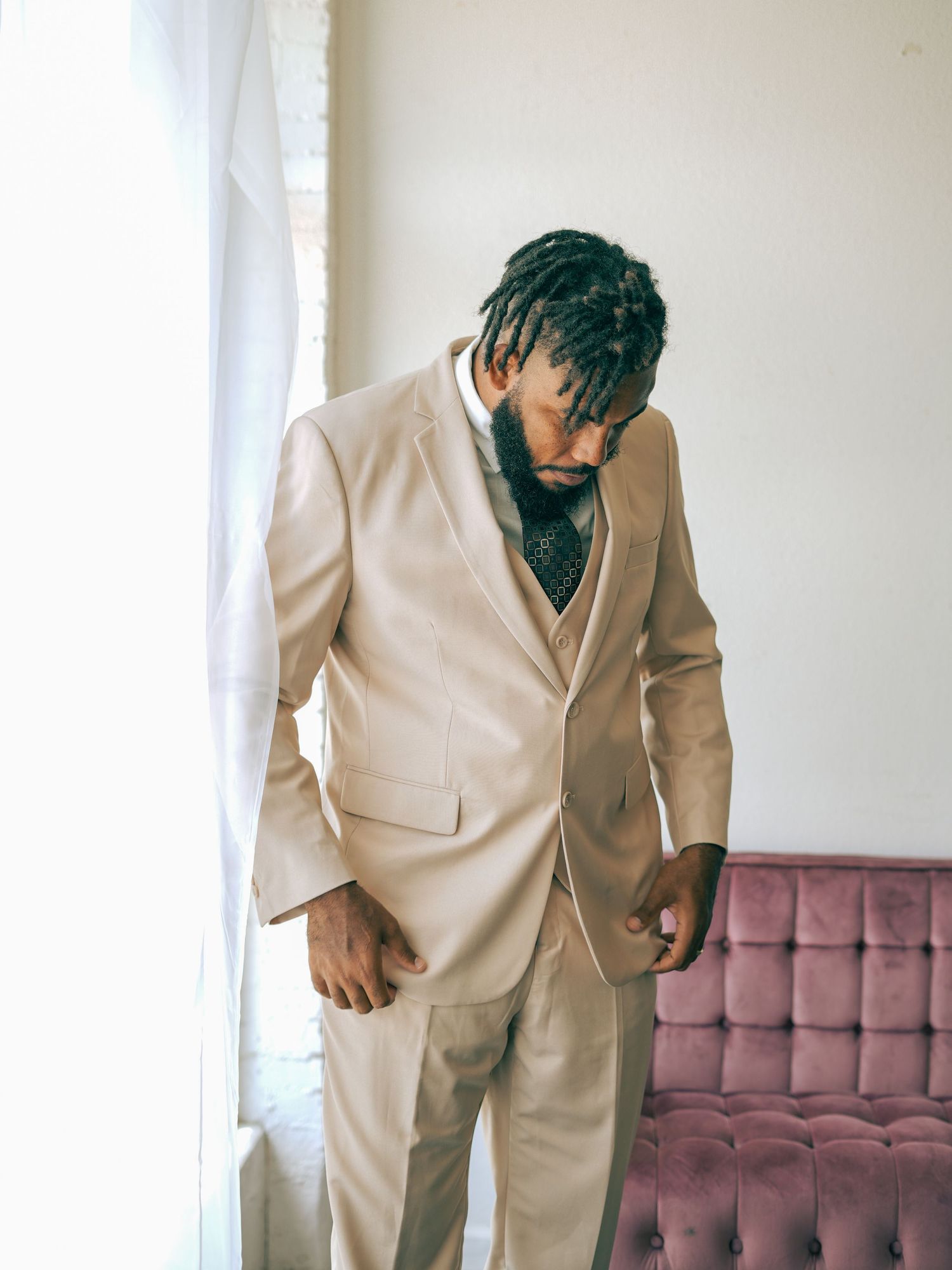 Man in beige suit looking down thoughtfully by window and pink couch.