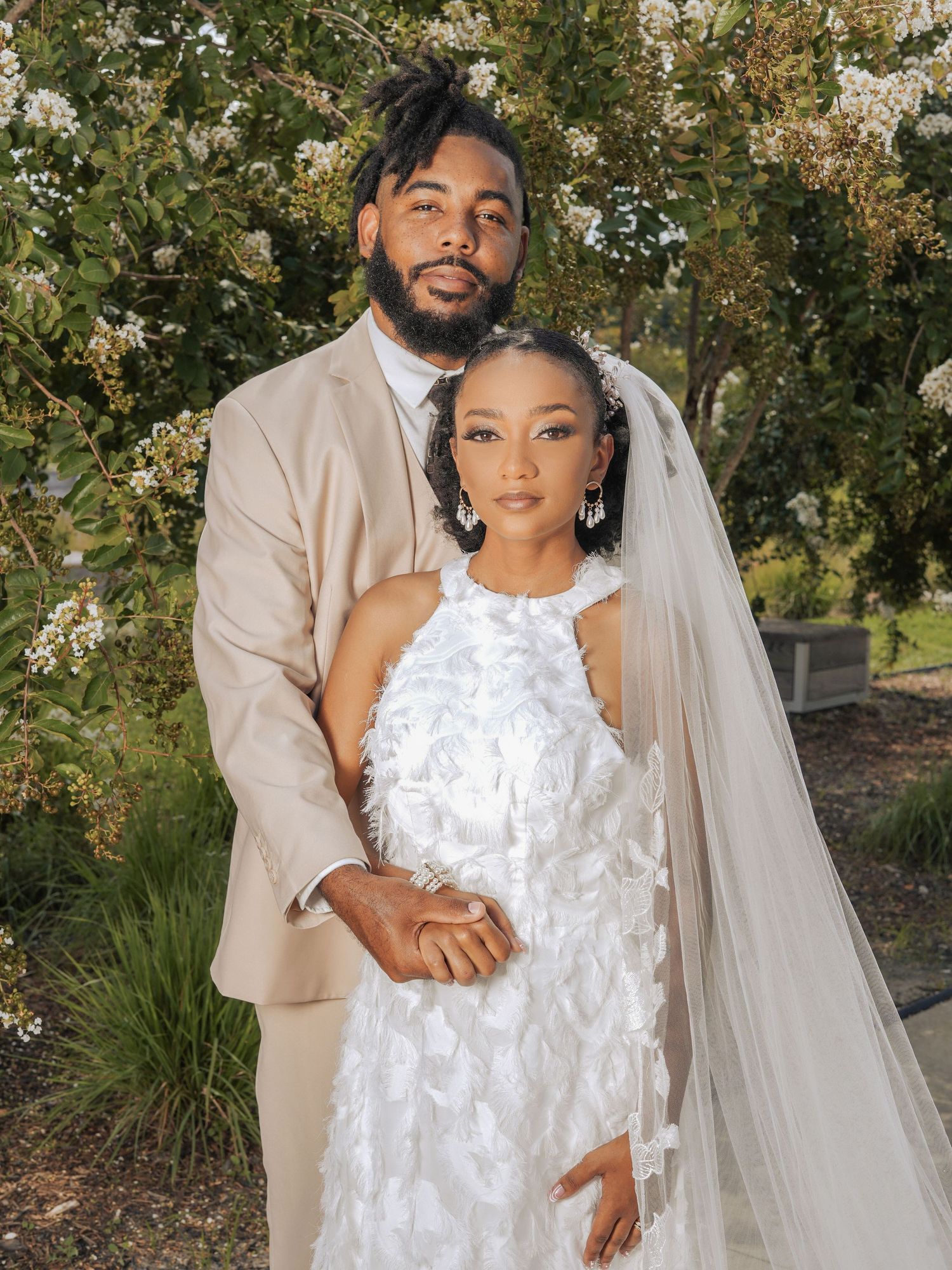 A wedding couple poses together outdoors against a backdrop of flowering trees.