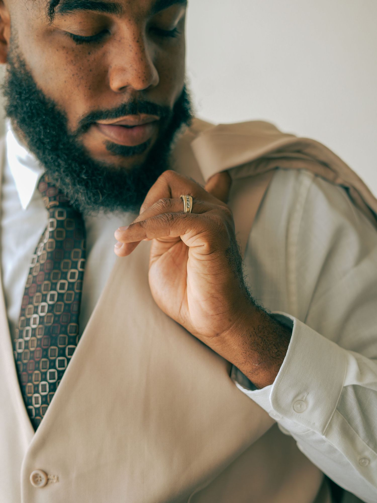 A groom in a beige vest and patterned tie adjusts his wedding ring.