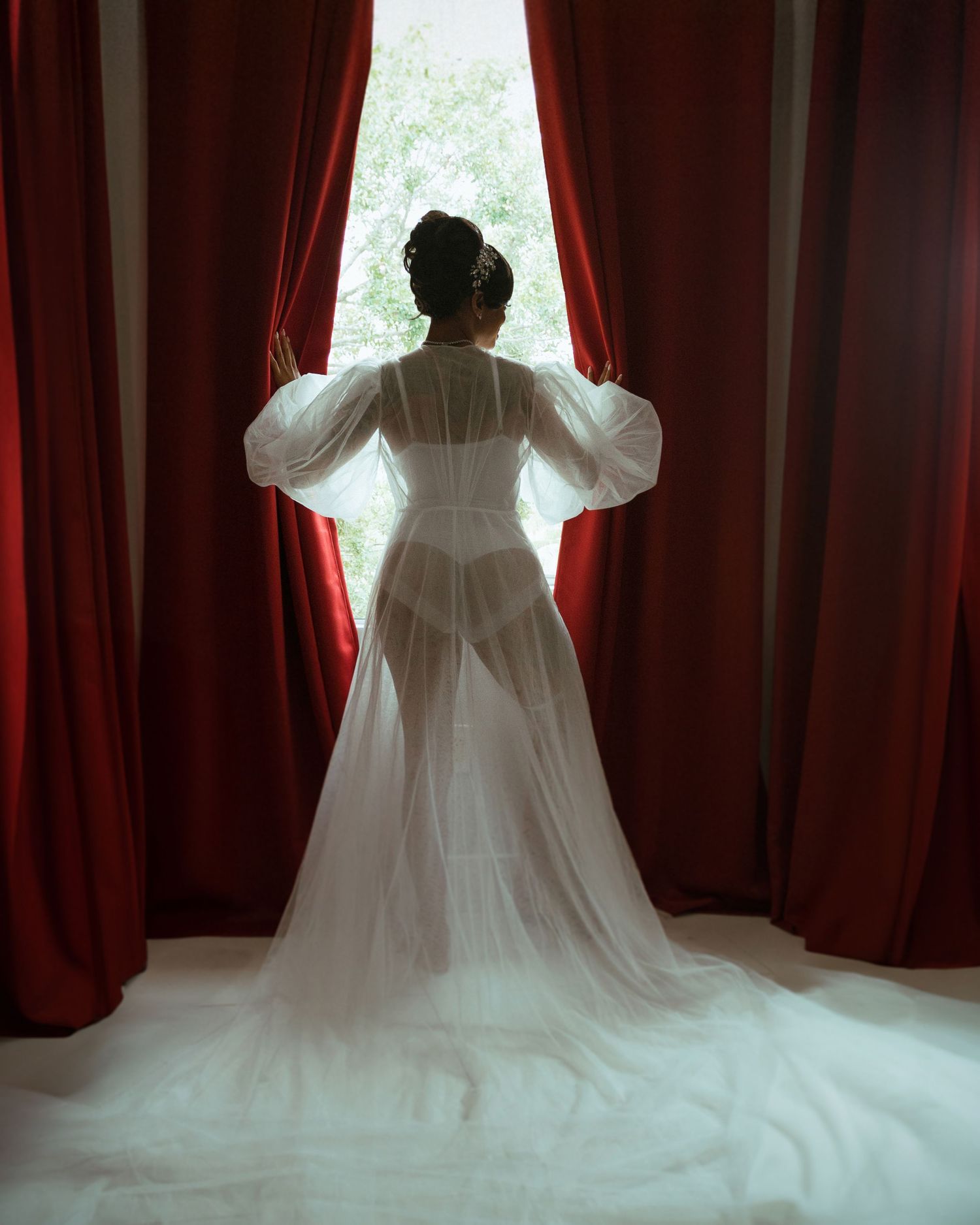 Silhouette of bride in flowing dress with dramatic red curtains and window light.
