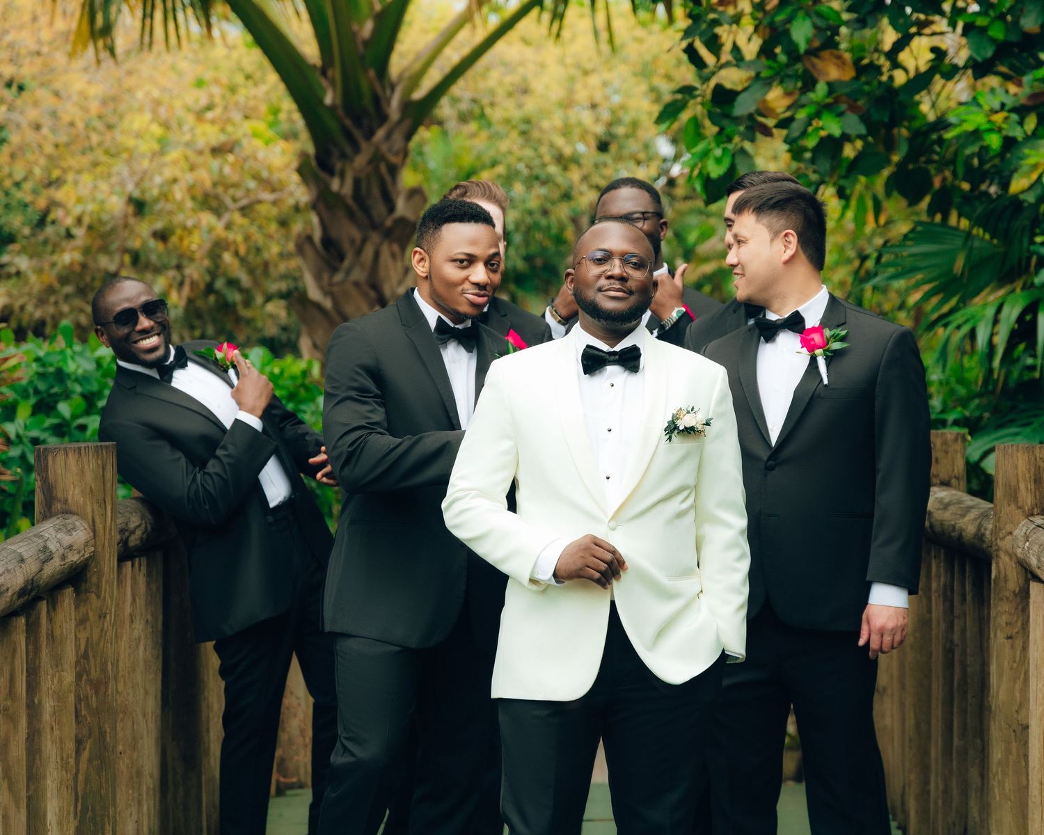 A group of groomsmen in formal attire laugh together in a lush garden setting during a wedding celebration.