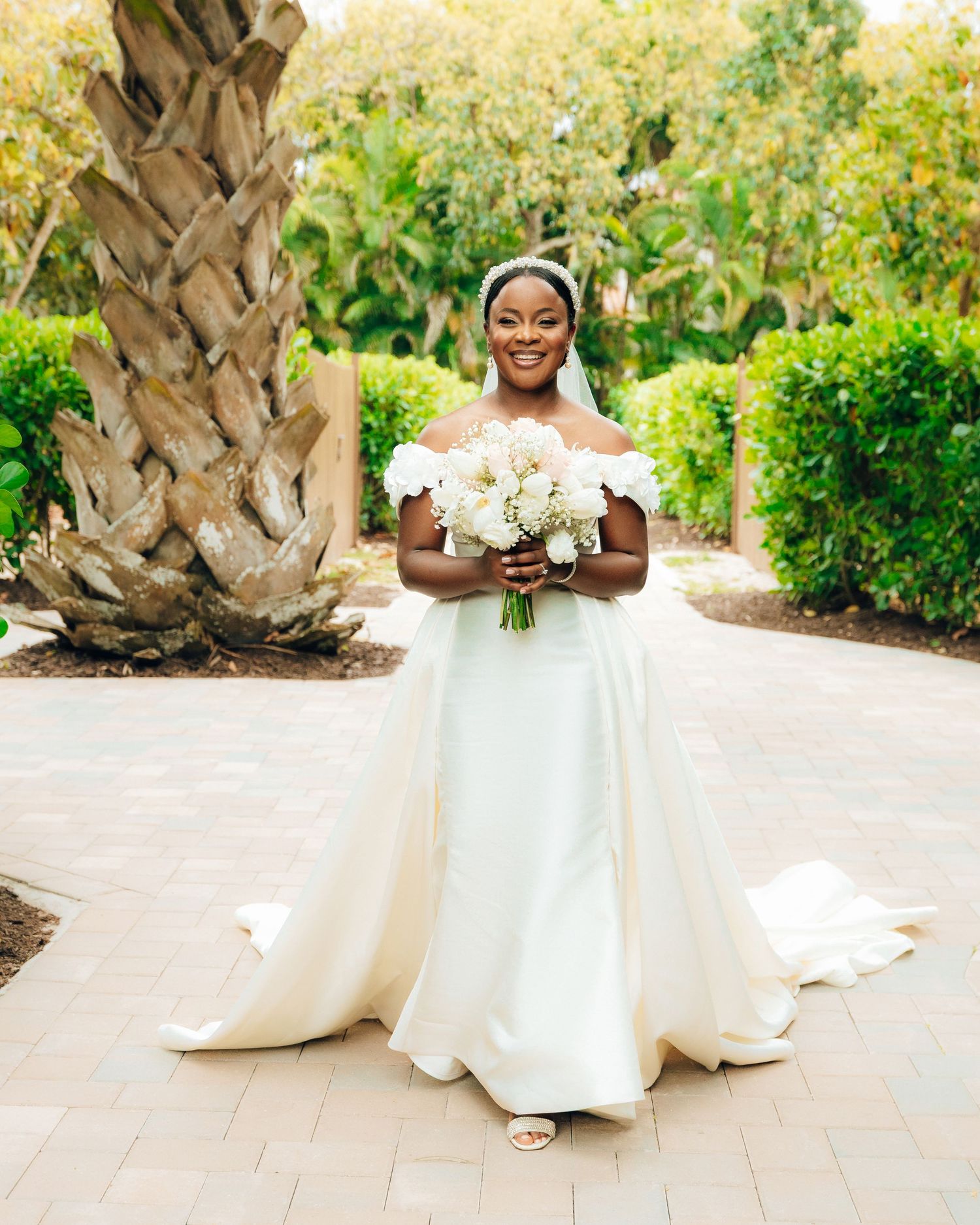 A bride in an elegant off-shoulder white wedding gown holds a bouquet while standing on a garden path near a palm tree.