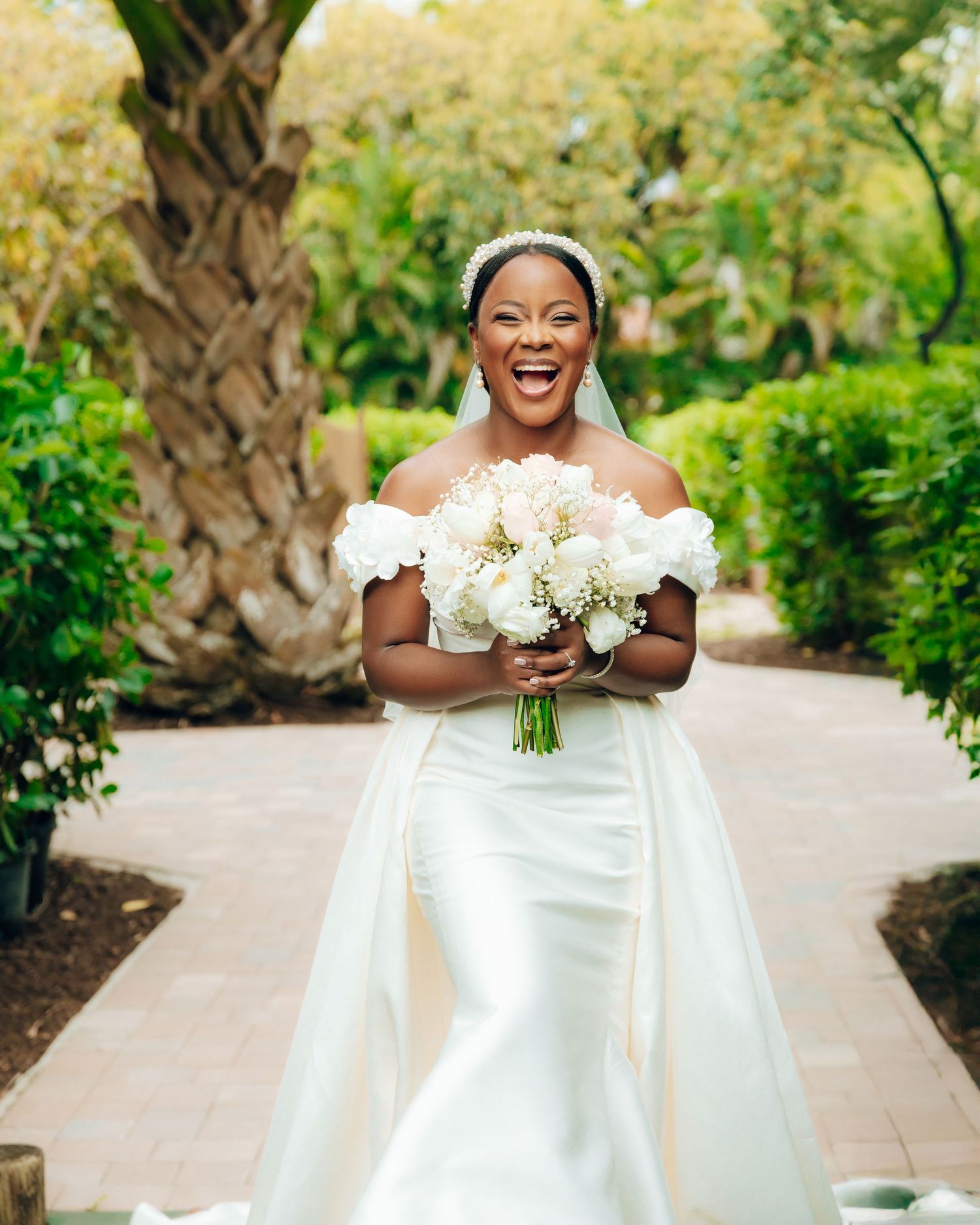 A joyful bride wearing an elegant off-shoulder white wedding gown holds a bouquet of white flowers in a garden setting.