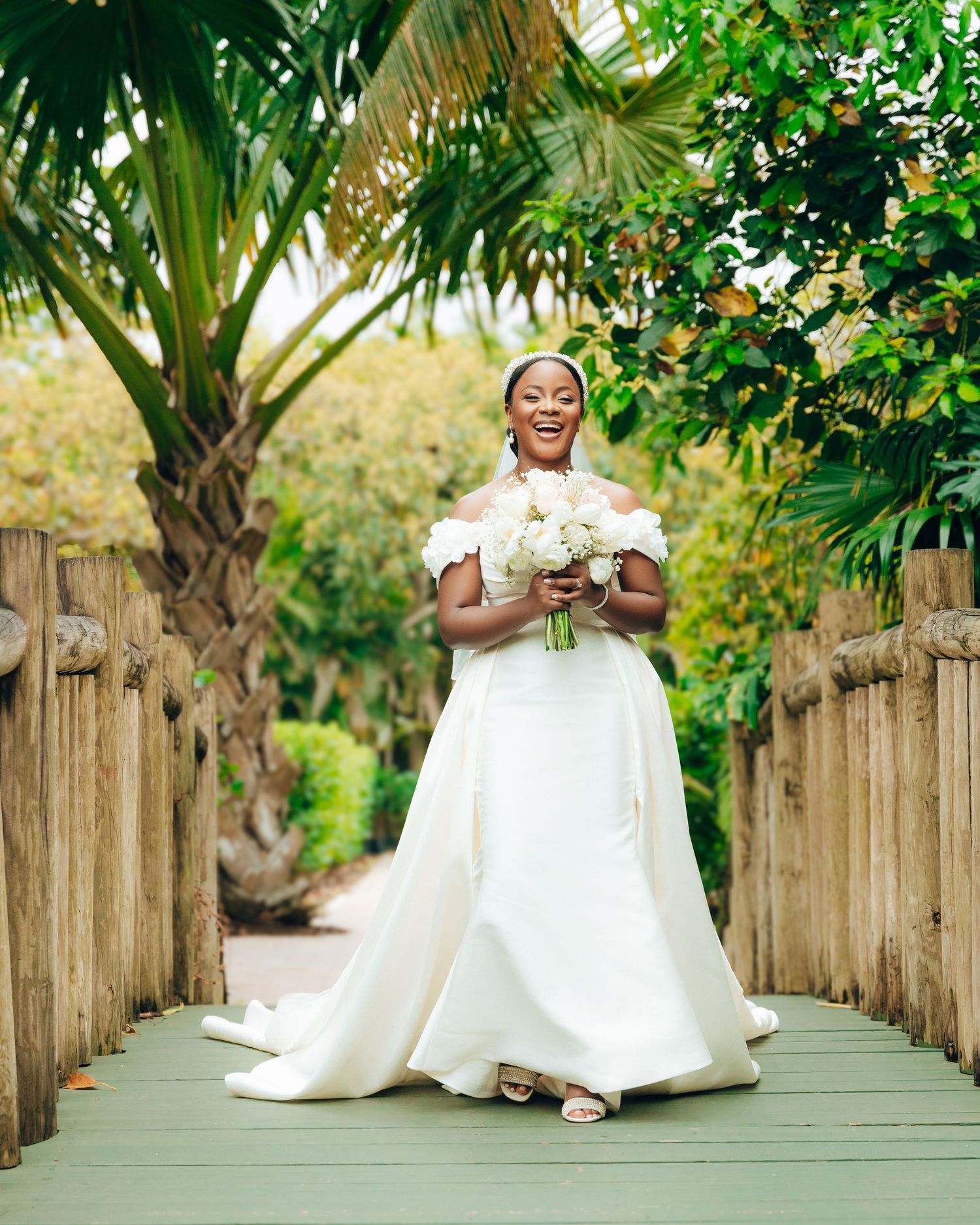 Someone in a stunning white off-shoulder wedding gown poses with a white bouquet on a wooden bridge surrounded by palm trees.