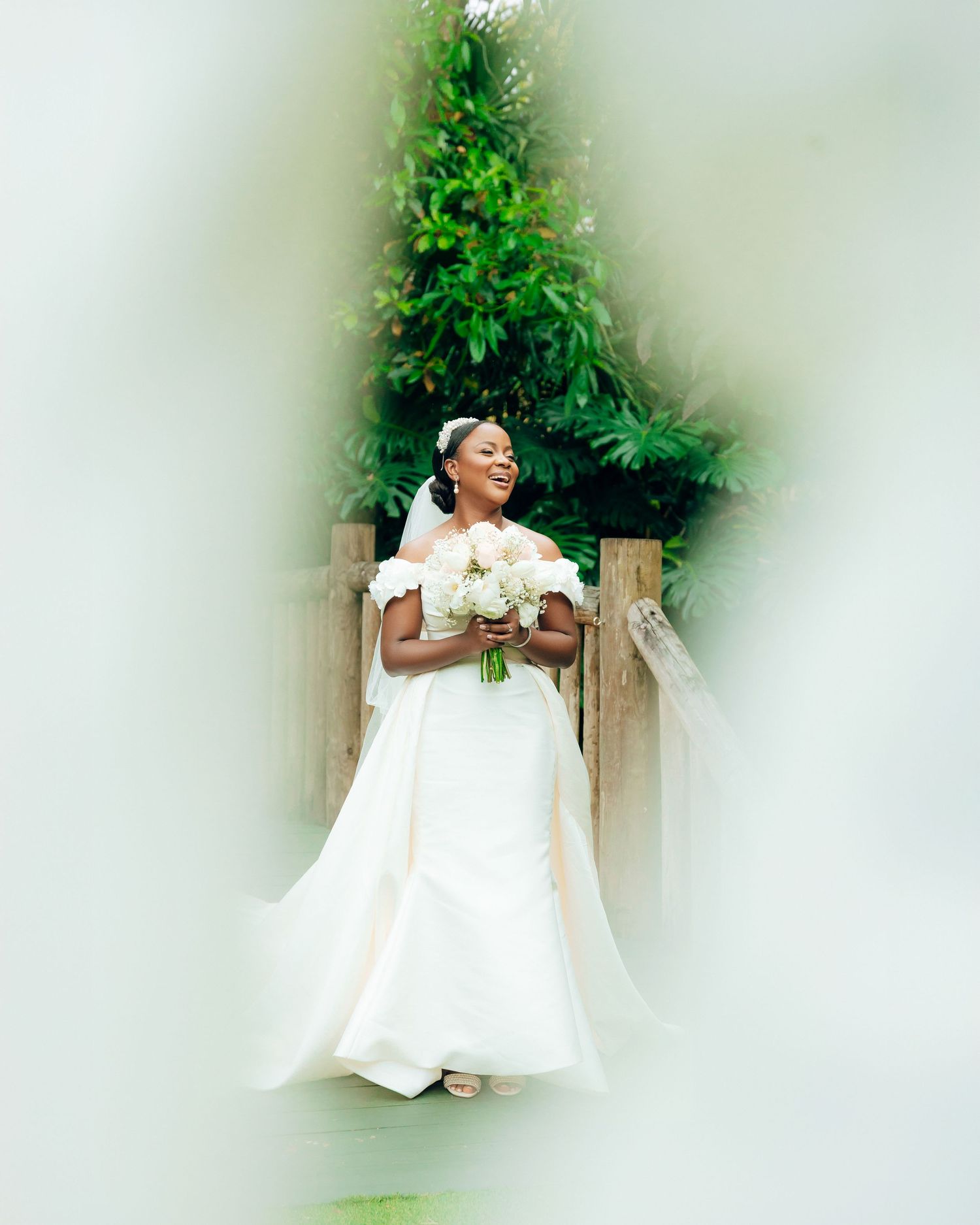 A joyful bride in an elegant white off-shoulder wedding dress holds a bouquet while standing against a garden backdrop.