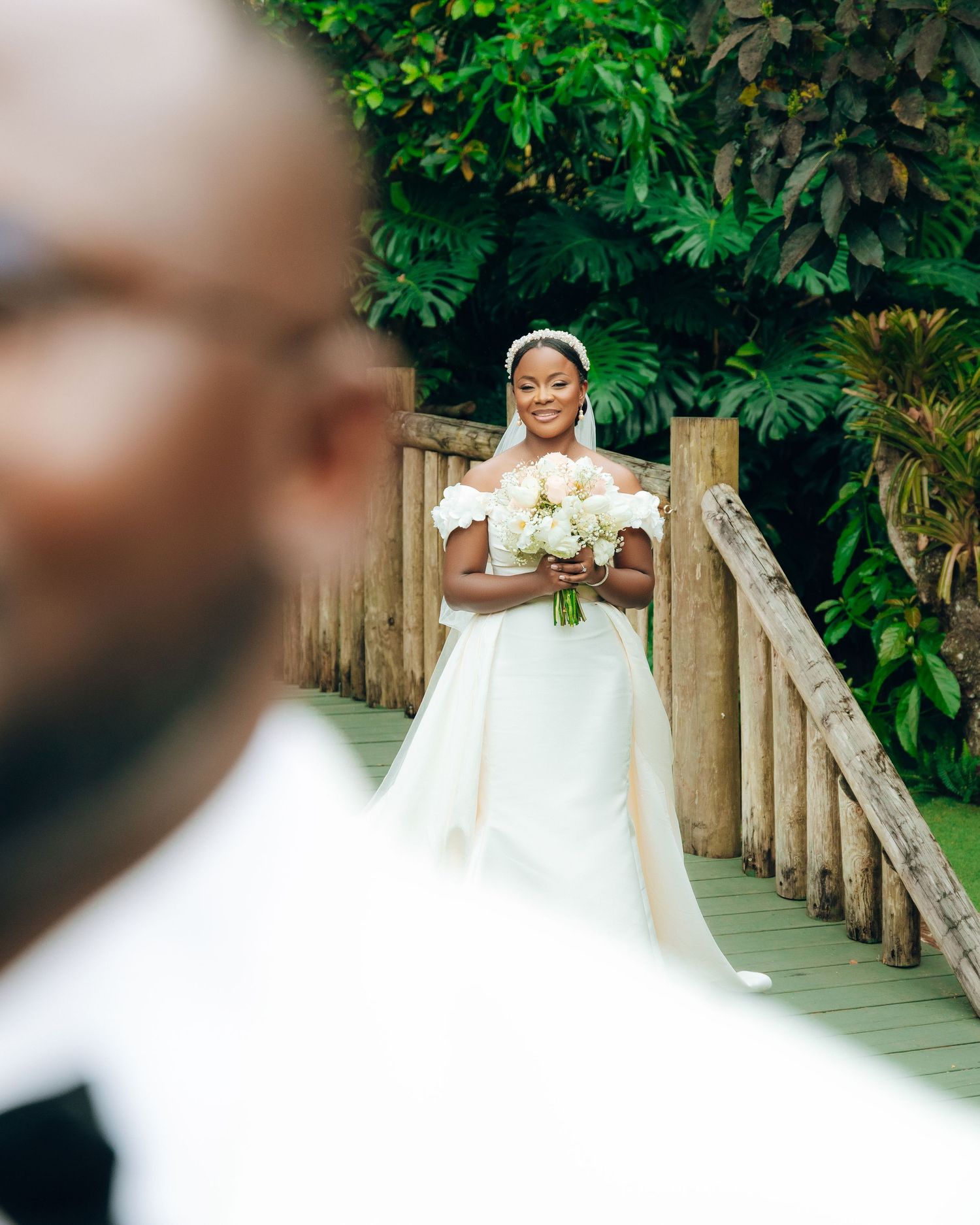 Beautiful bride in an off-shoulder white gown holds a bouquet on a wooden bridge surrounded by lush tropical foliage.
