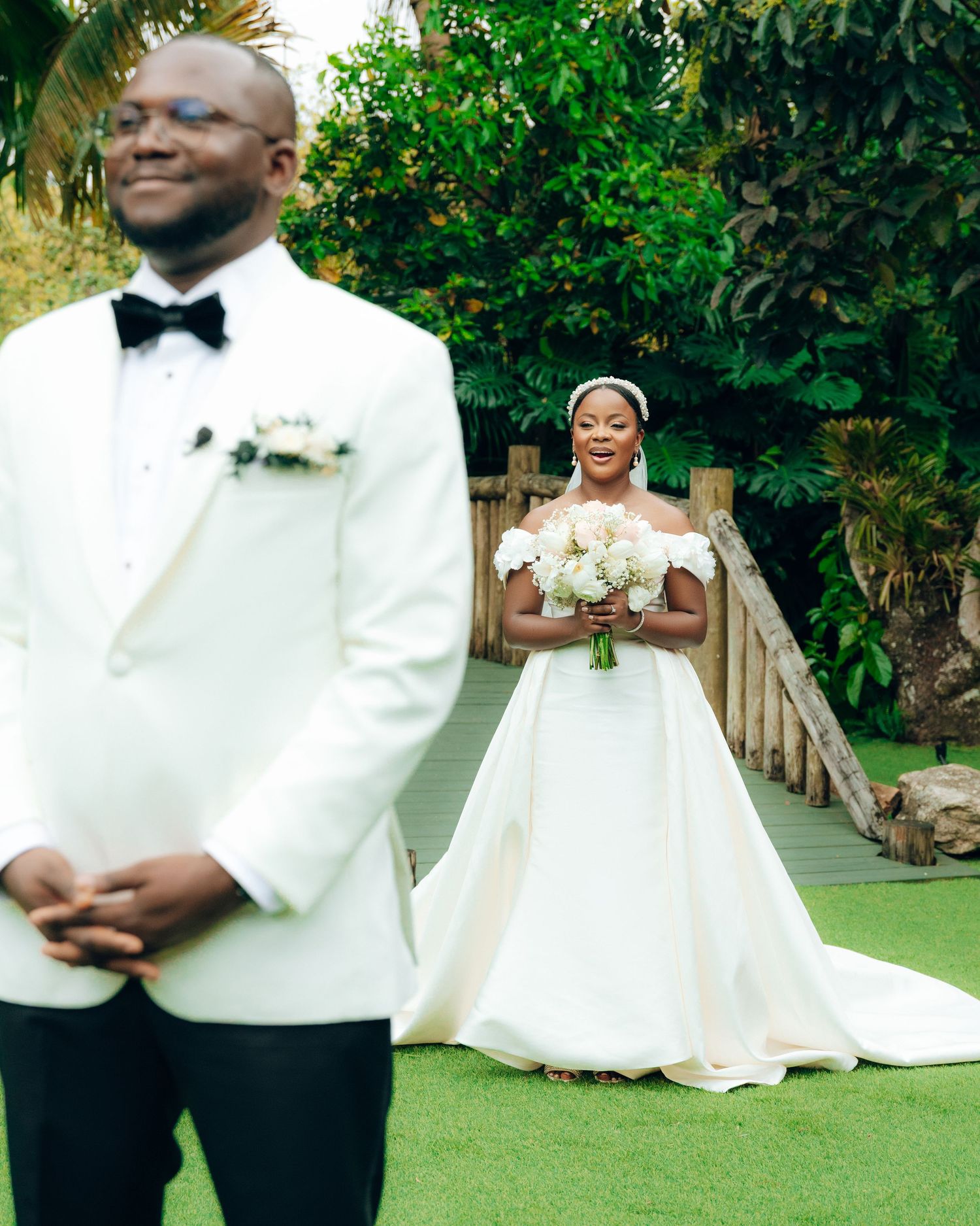 A wedding first look moment on a garden path with the bride holding a white bouquet and groom in white tuxedo jacket.