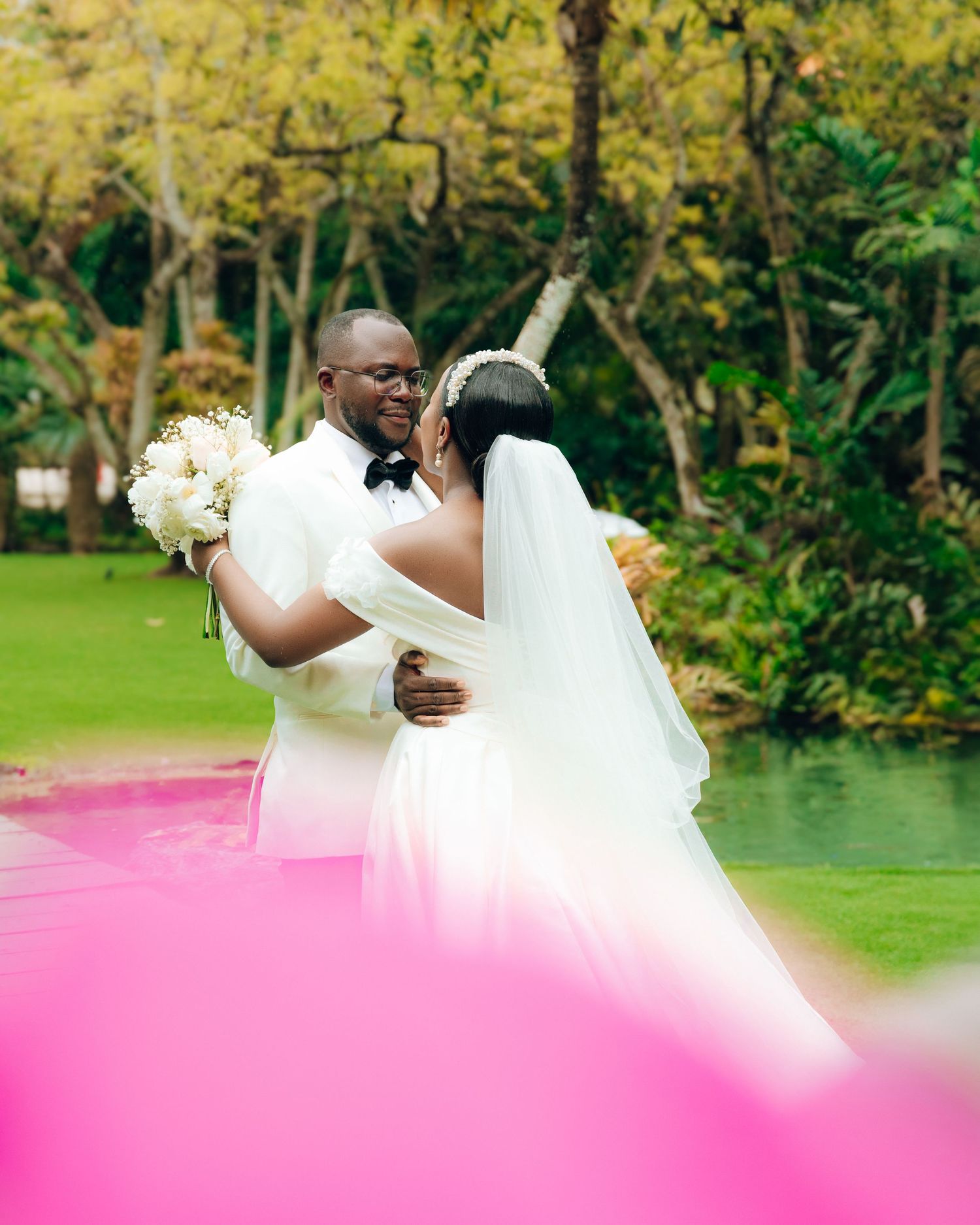 Two people share a romantic moment in white wedding attire by a pond surrounded by autumn trees in a garden setting.