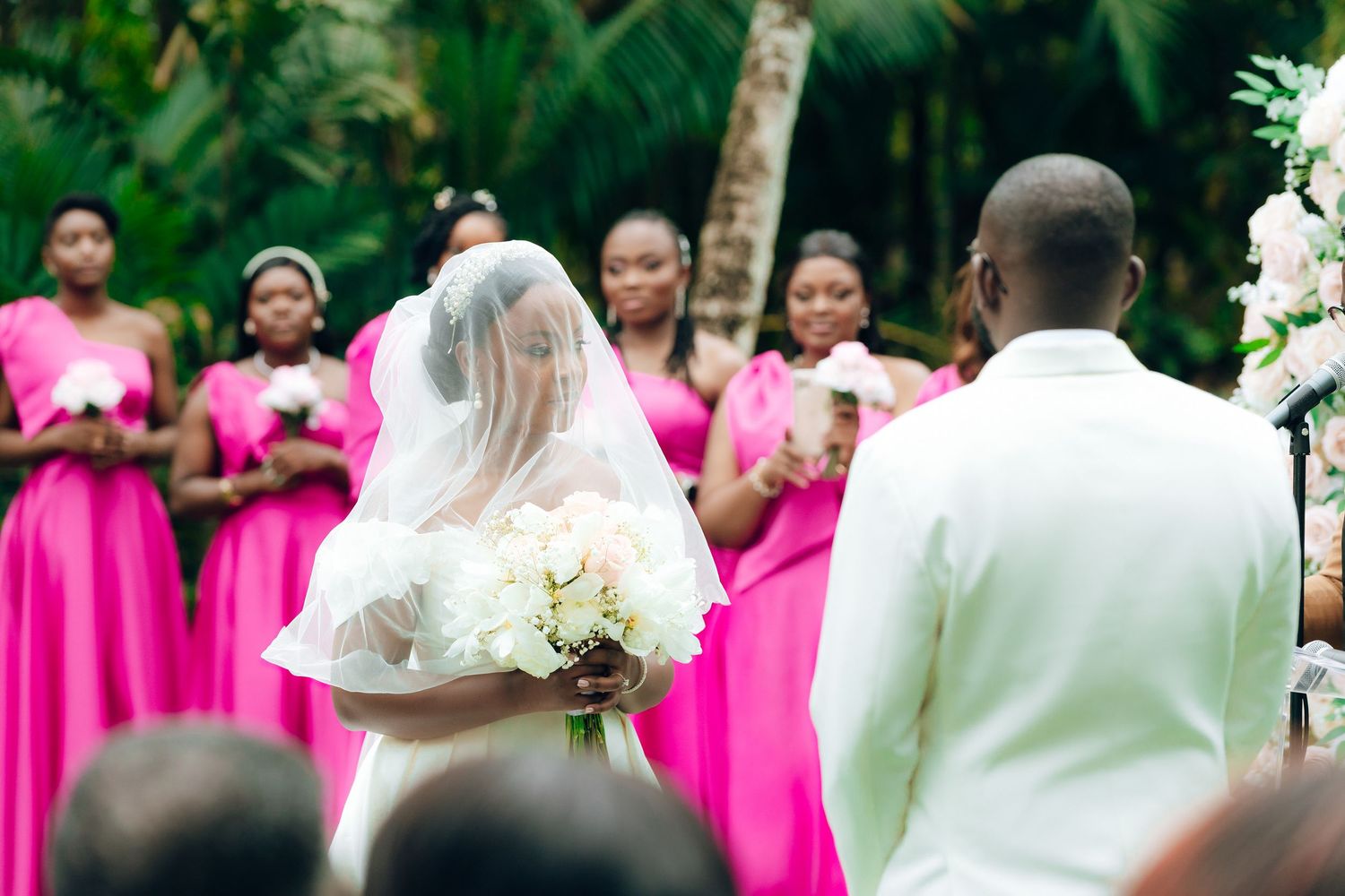 Bride in white veil and bridesmaids wearing bright pink dresses stand during outdoor wedding ceremony.