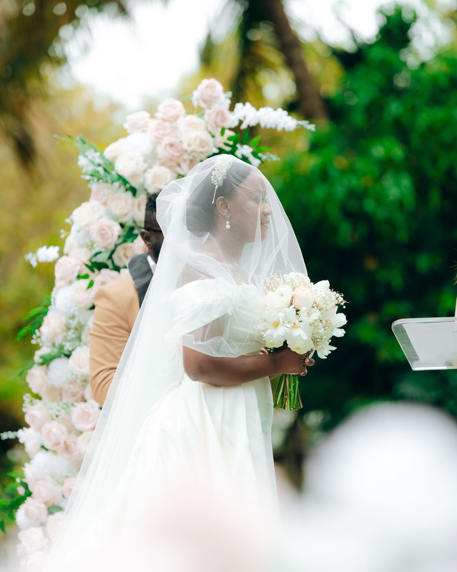 A bride in a white wedding dress and veil holds a bouquet beneath an elegant floral arch during an outdoor ceremony.