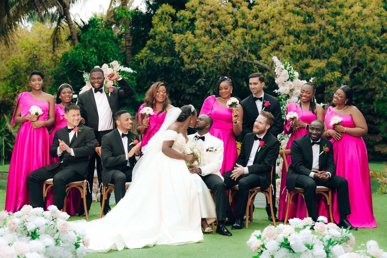 Wedding party in bright pink bridesmaid dresses and black tuxedos pose together outdoors with floral decorations.
