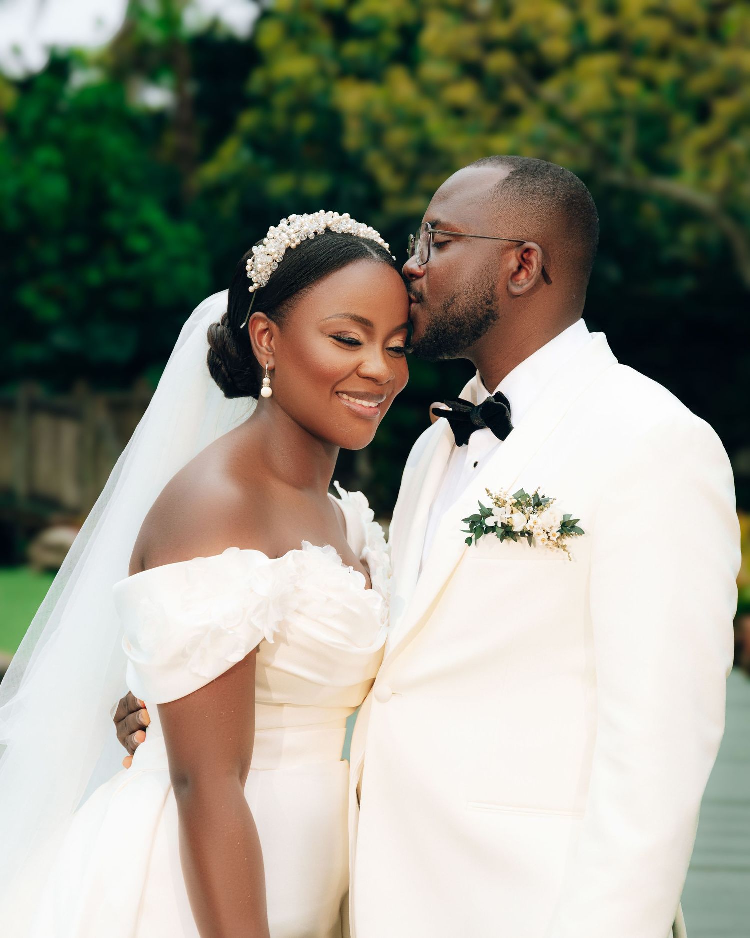 A joyful newlywed couple share an intimate moment outdoors, wearing white wedding attire with beautiful green foliage.