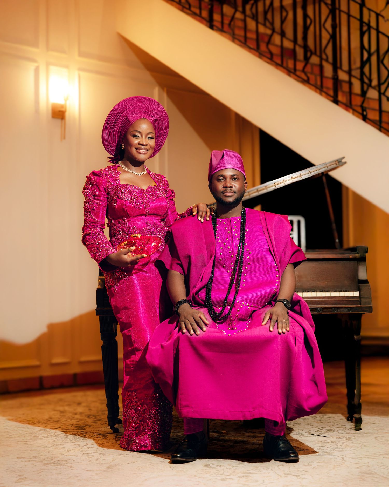 A couple in vibrant fuchsia traditional Nigerian attire pose together by a grand piano.
