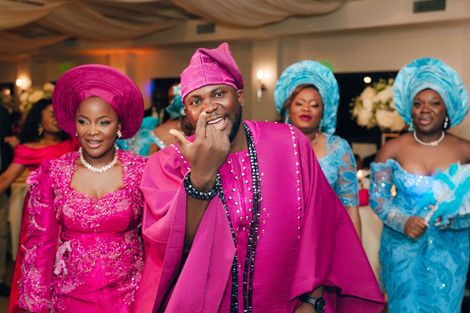 A group wearing vibrant pink and blue traditional African formal attire with elaborate head wraps at a celebration.