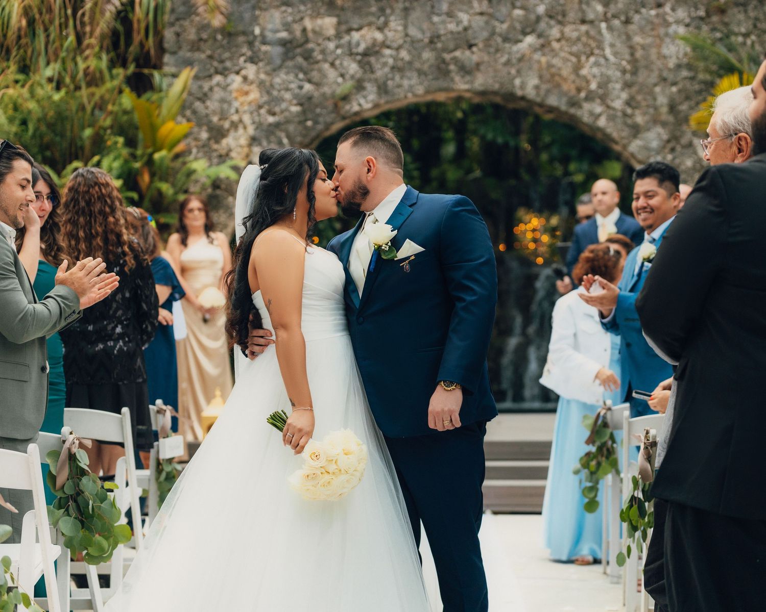 A romantic wedding ceremony moment under a stone archway surrounded by guests and tropical foliage.
