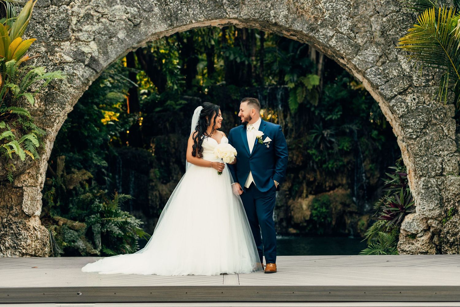 A wedding couple shares an intimate moment under a rustic stone archway in a garden setting.