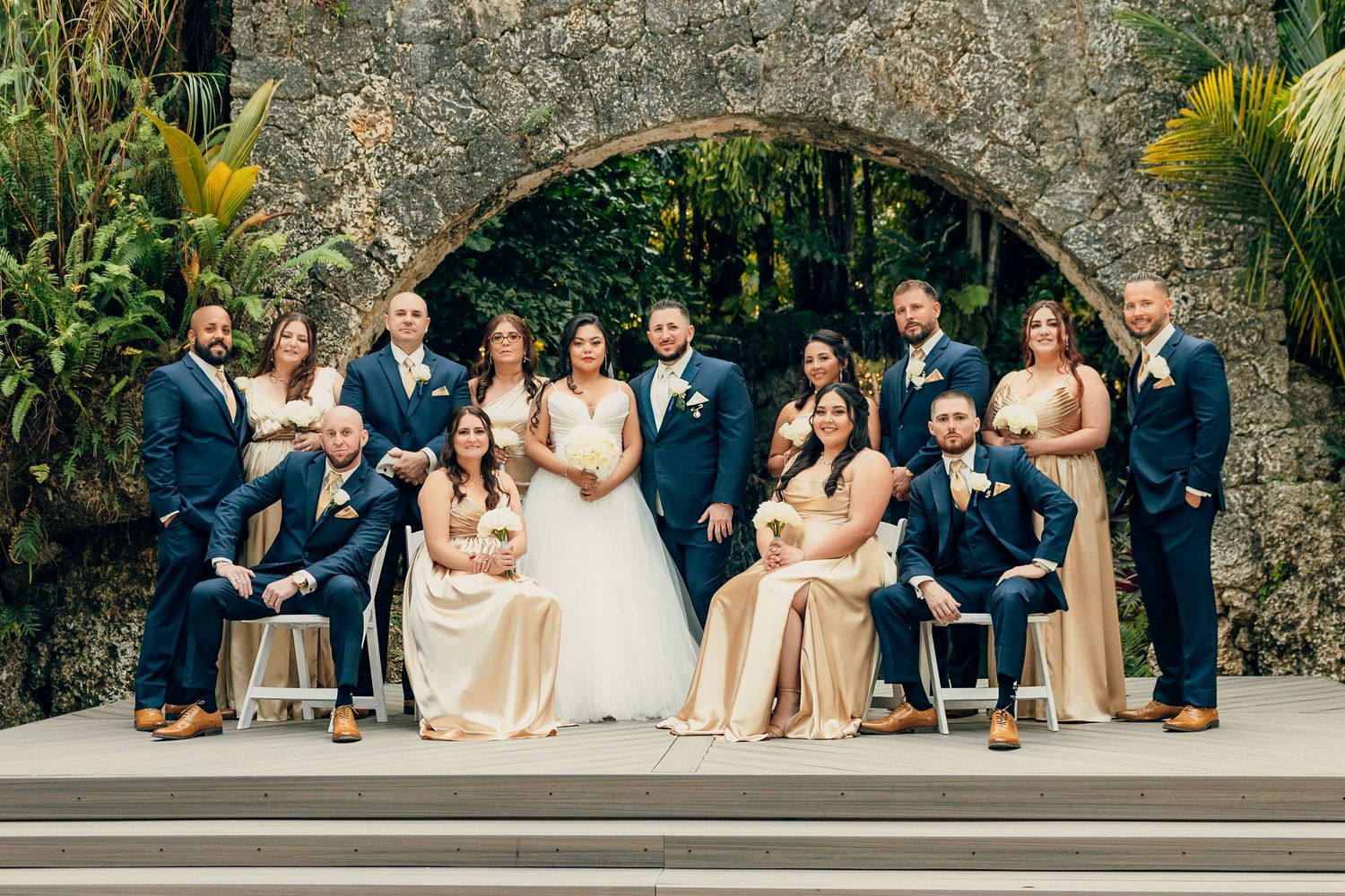 Wedding party in navy and champagne attire poses together under a stone archway.