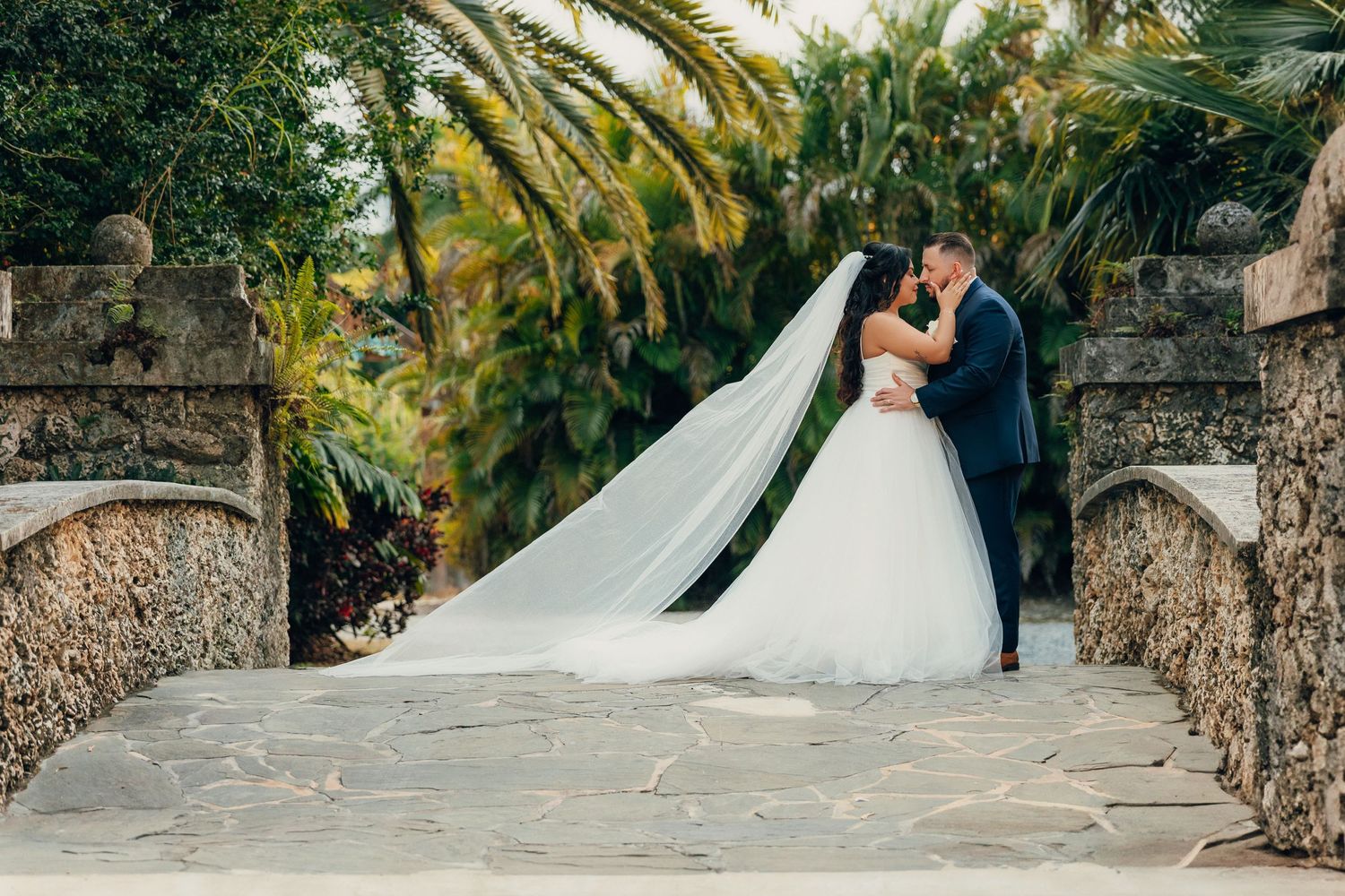A wedding couple embraces on a stone pathway surrounded by lush tropical gardens.