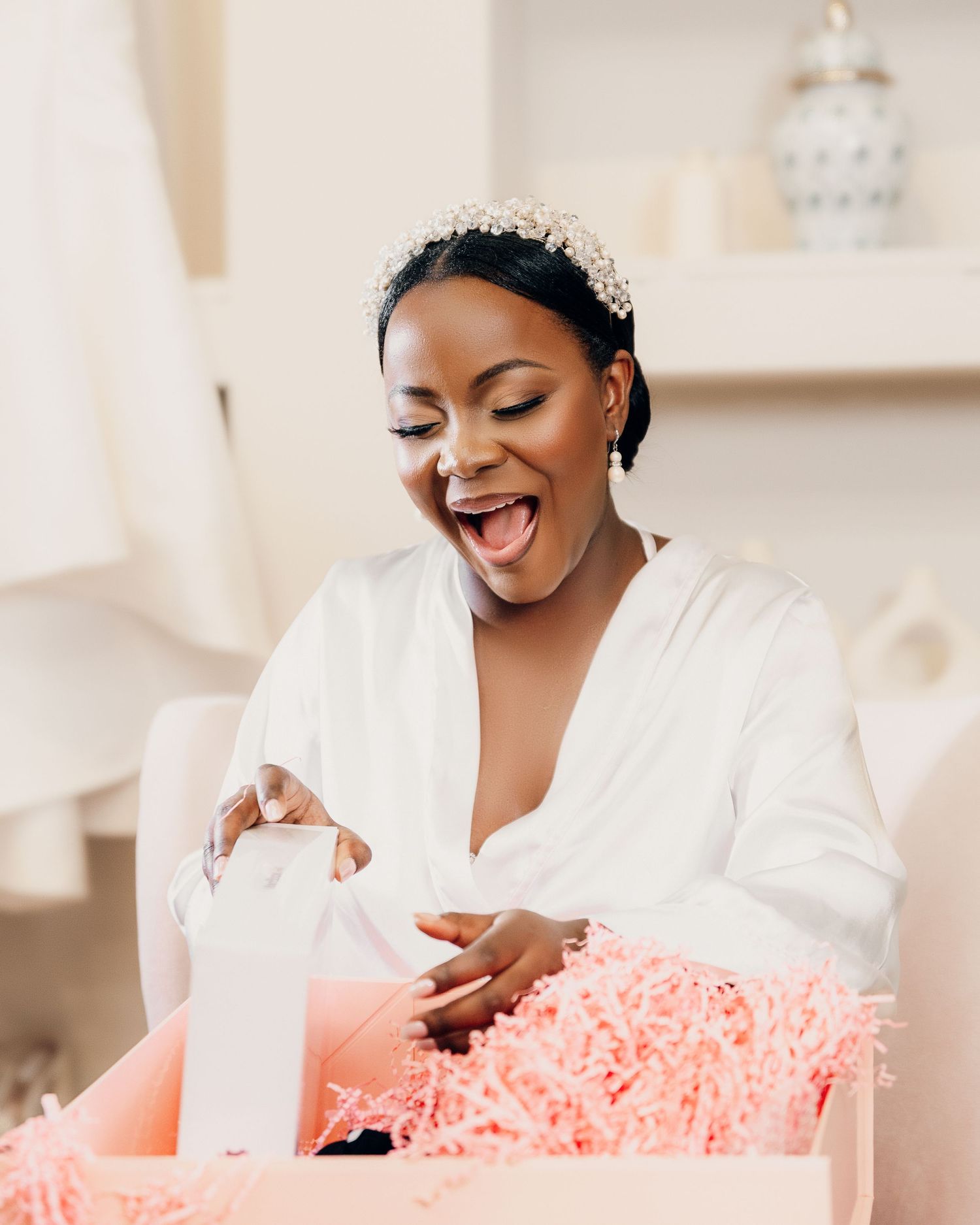 A joyful woman in a white robe opens a gift box with pink tissue paper while getting ready for a special occasion.