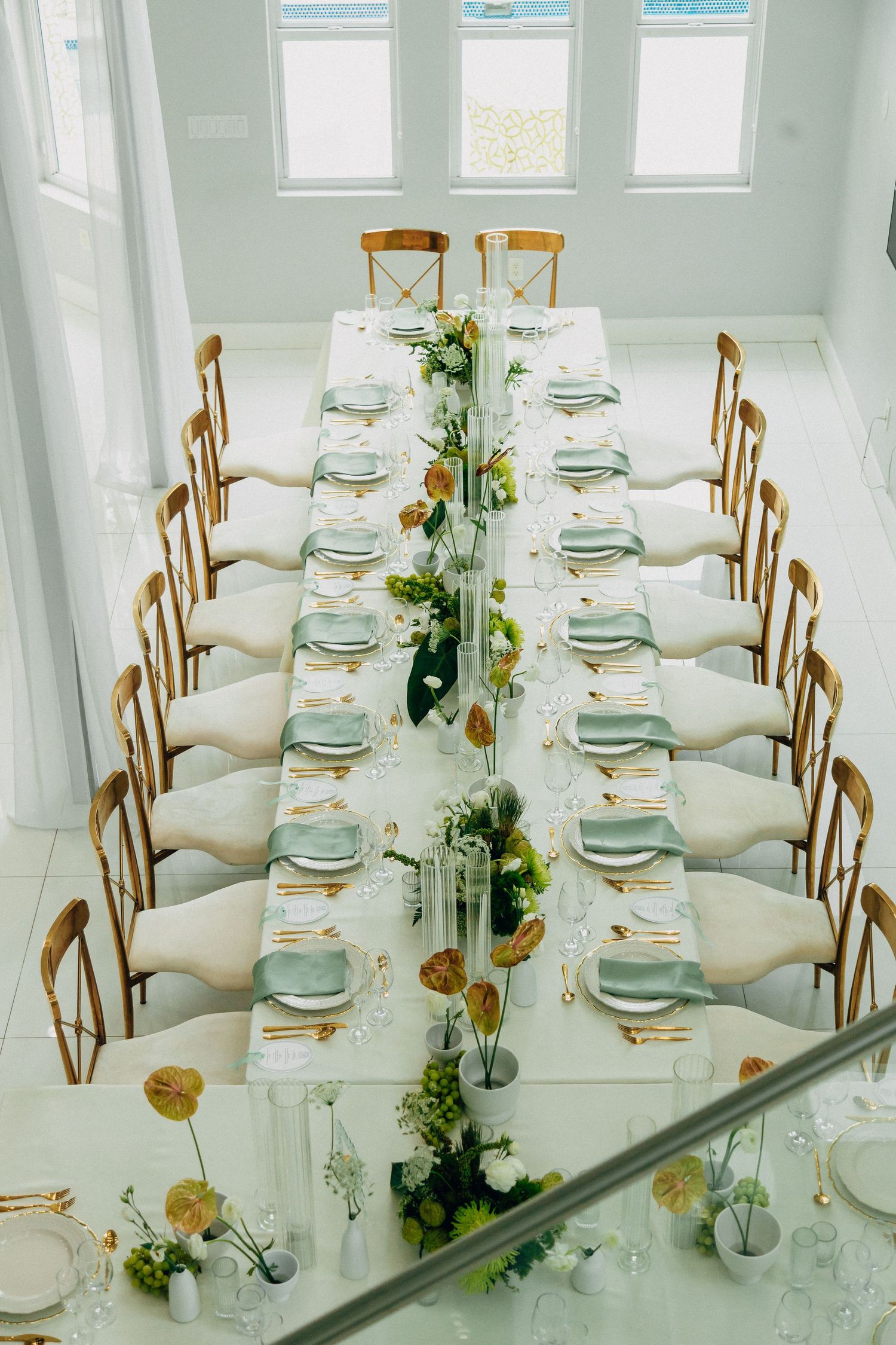 Elegant long wedding table setting with wooden chairs and delicate floral centerpieces in a bright white room.