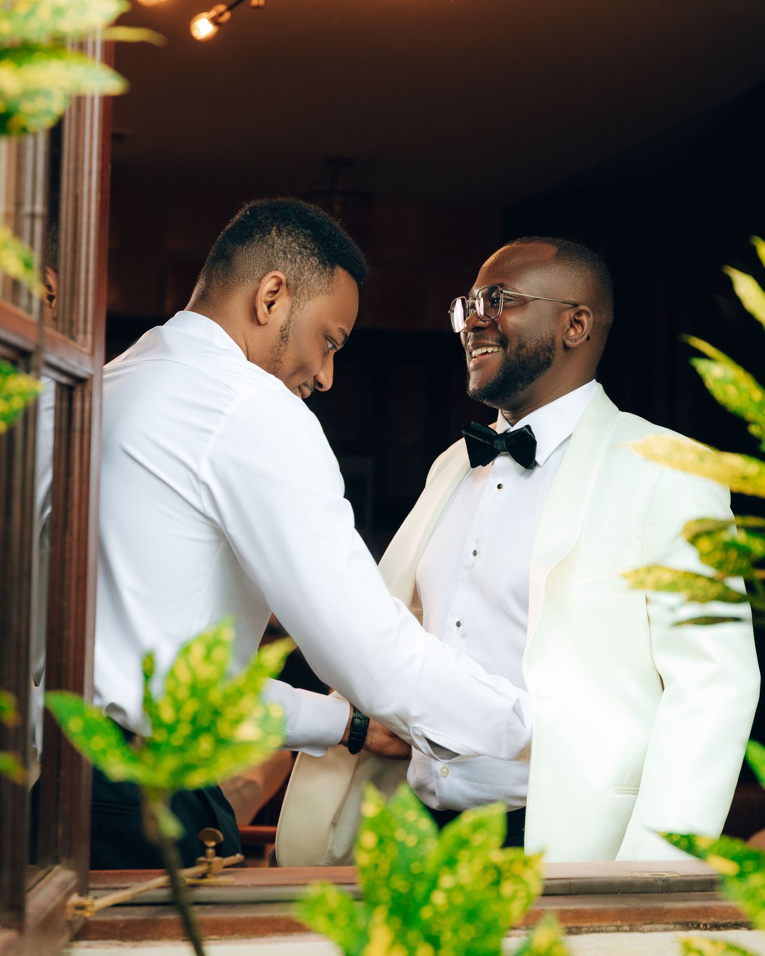 Two people in white tuxedos share a joyful moment while getting ready for a formal event.